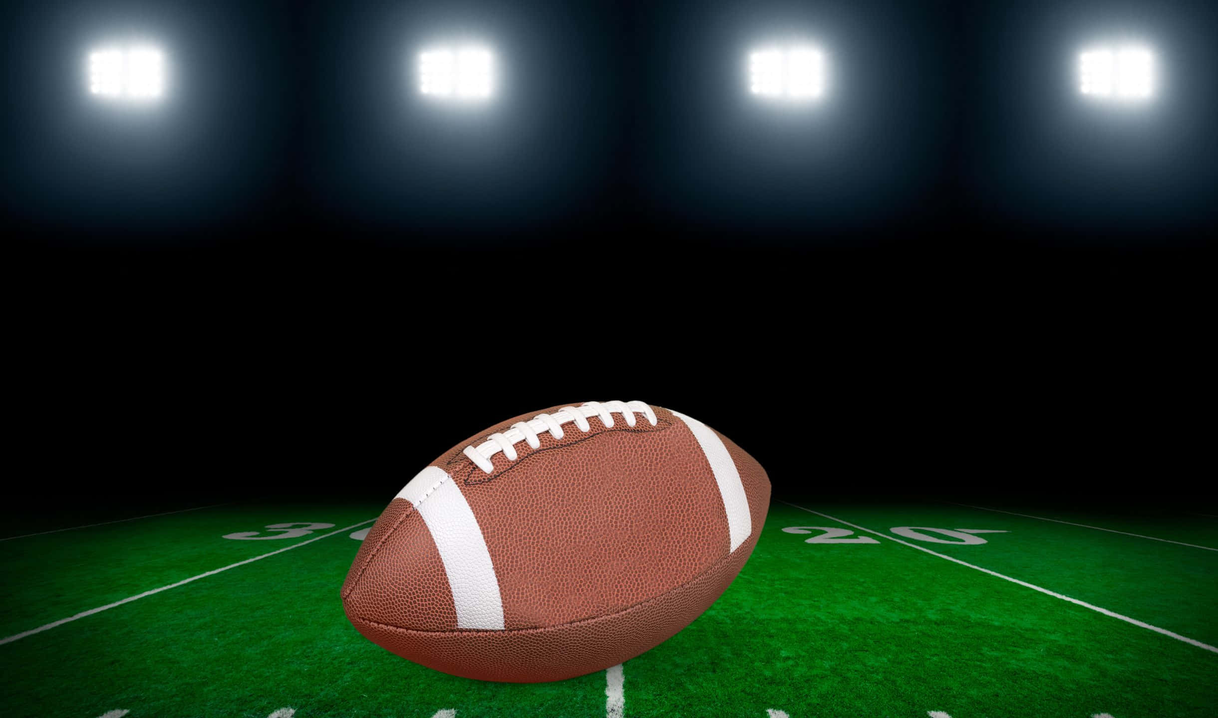 2440x1440 Football 桌布 壁紙 — 一個棕色皮革足球在夜間明亮的體育場燈光下放在綠色場地上。