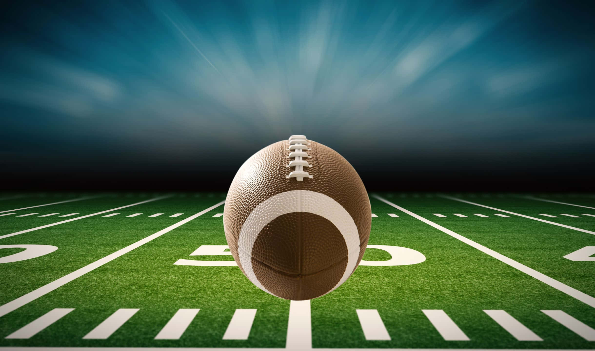 2440x1440 Football 桌布 壁紙 — 一個棕色美式足球居中放置在充滿活力的綠色球場上，球場有白色碼線，上方是戲劇性的藍天，帶有耀眼的光束。