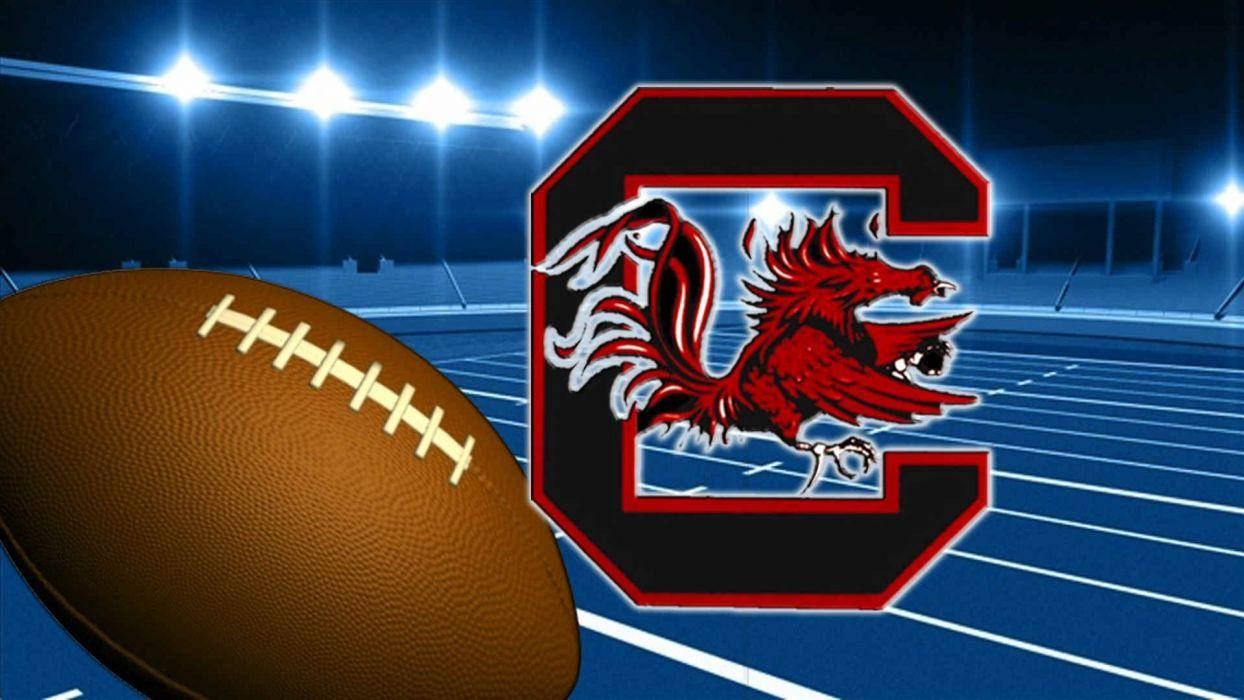 South Carolina Gamecocks 桌布 壁紙 — 一個棕色美式足球放在藍色球場上，在明亮的體育場燈光下，旁邊有一個紅色和黑色的遊戲標誌，上面有一隻兇猛的公雞。