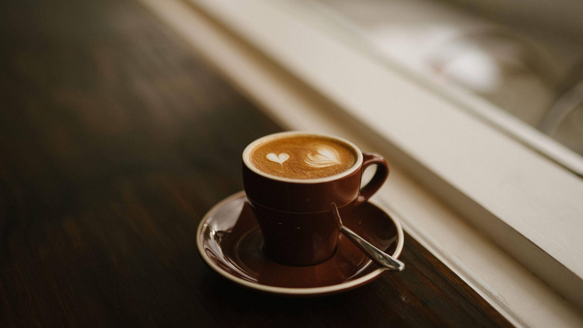 Morning 桌布 壁紙 — 一個棕色陶瓷杯裝的拿鐵，帶有心形奶泡，放在碟子上，置於陽光照射的窗台上。