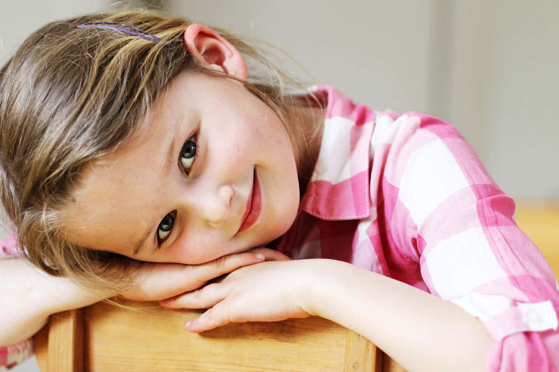 kid 桌布 壁紙 — 一個棕色頭髮、綠色眼睛的女孩，將下巴擱在木質表面上的手上，溫暖地微笑。