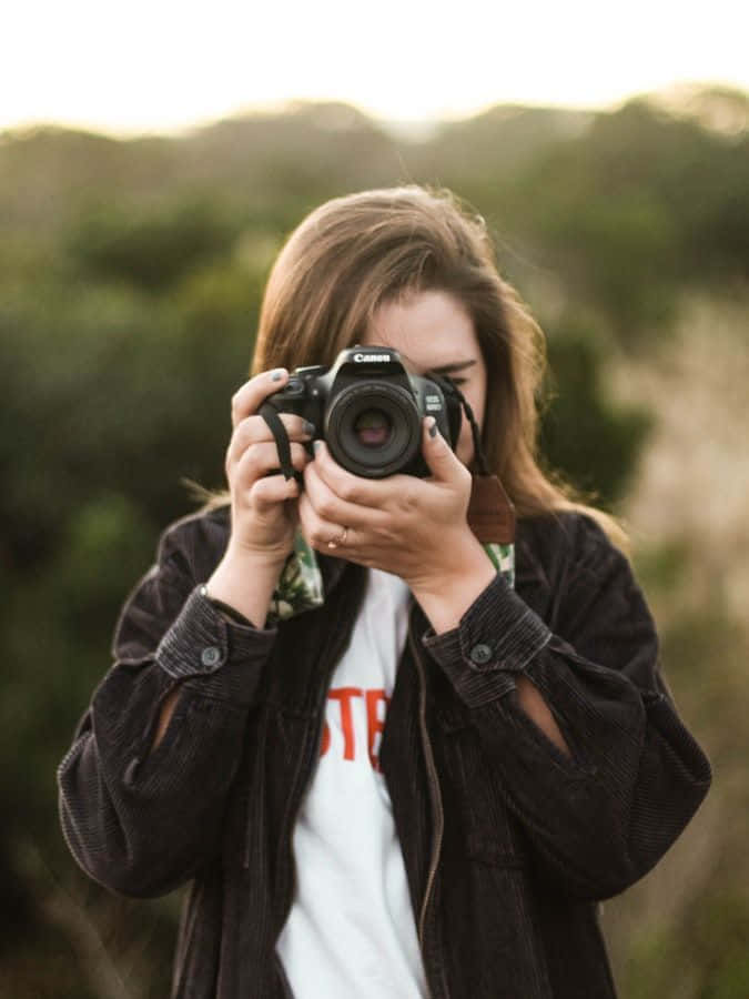 Holding Camera 桌布 壁紙 — 一個棕色頭髮的人將黑色相機舉到臉前，背景是柔和模糊的綠色和米色戶外背景。