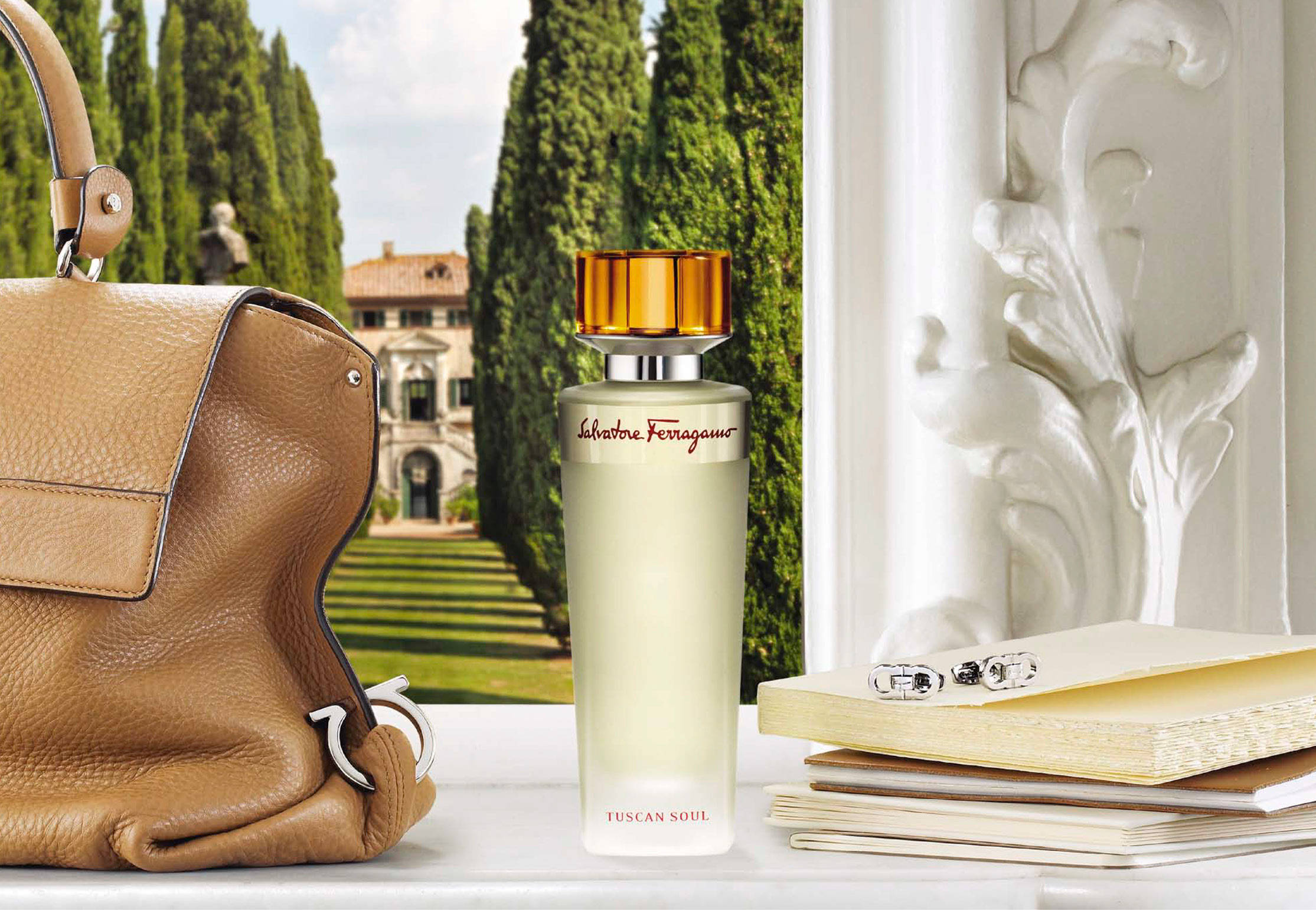 Ferragamo 桌布 壁紙 — 一個棕褐色皮革手提包、香水瓶和筆記本放在大理石窗台上，俯瞰著被高大柏樹環繞的托斯卡納別墅。