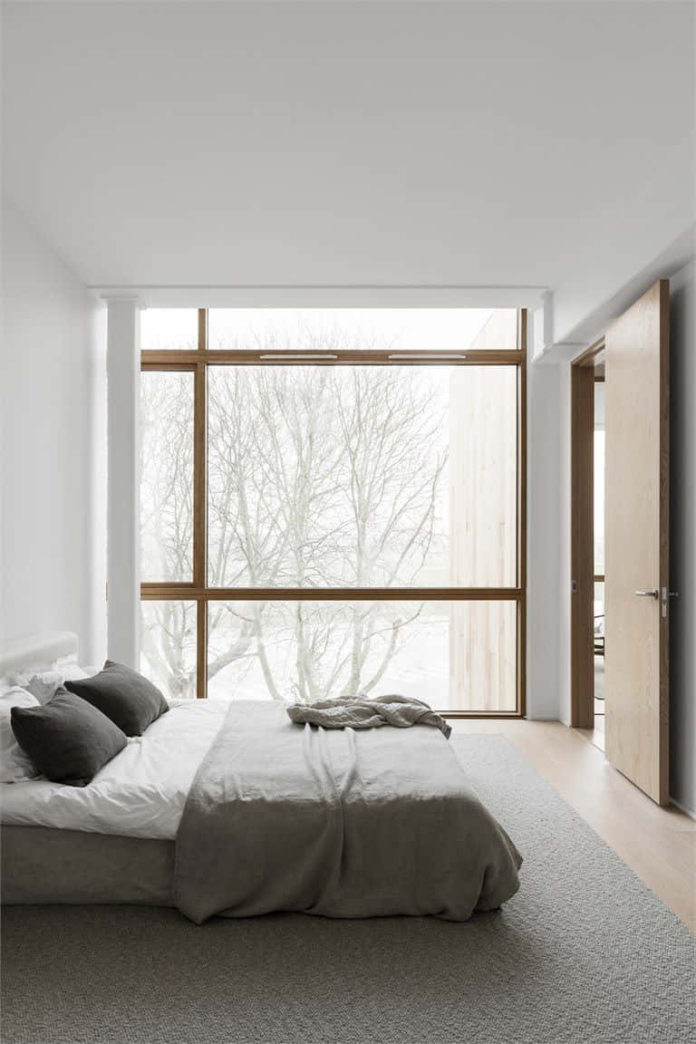 Minimalist Design 桌布 壁紙 — 一個極簡主義的臥室，大窗戶框出外面光禿禿的冬日樹木，柔和自然光，一張鋪著灰色床單的整齊床鋪。