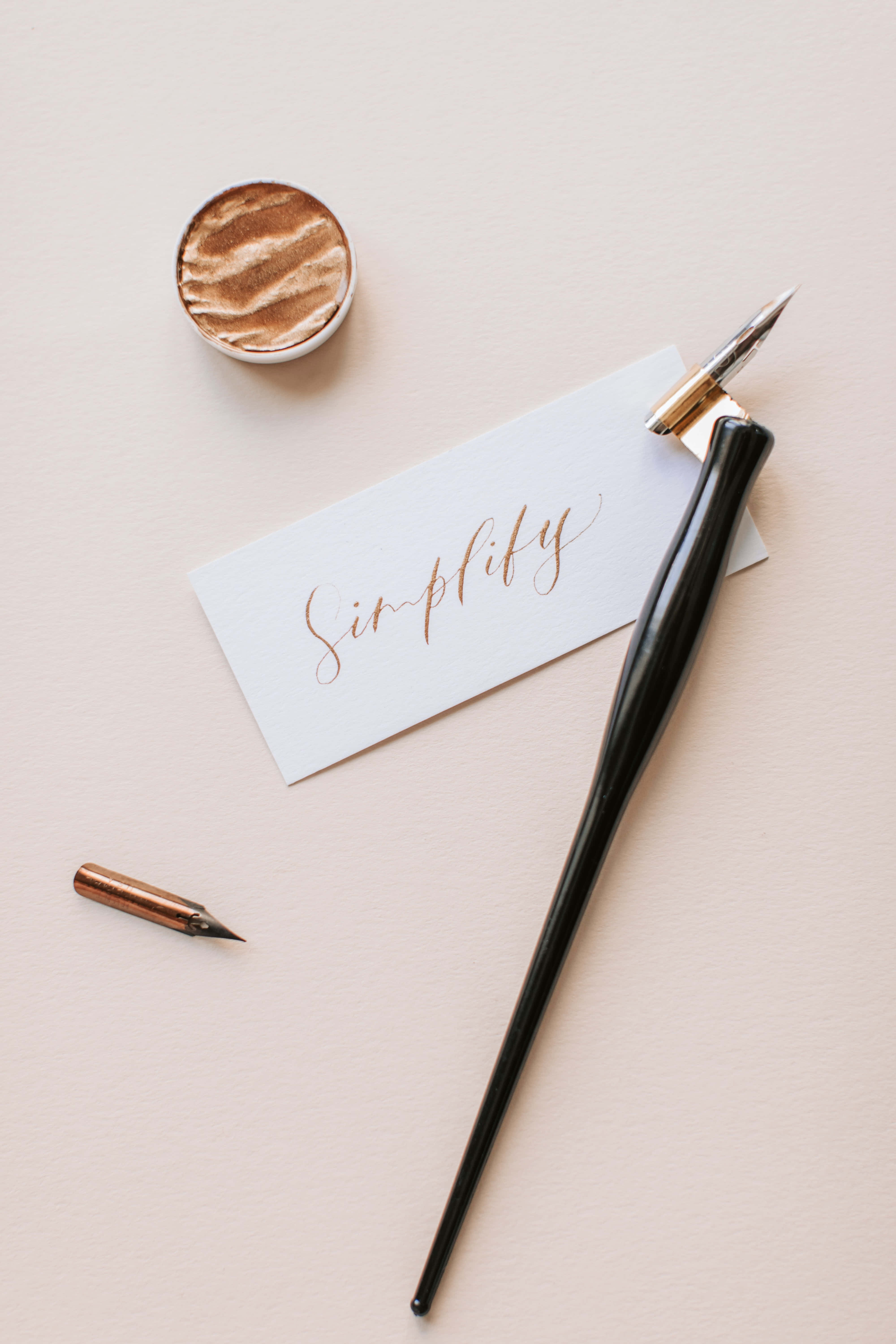 calligraphy 桌布 壁紙 — 一個極簡的平鋪構圖，黑色金筆尖的鋼筆放在一張帶有金色書法字 simplify 的卡片和一個小墨水瓶上，背景是柔和的米色。