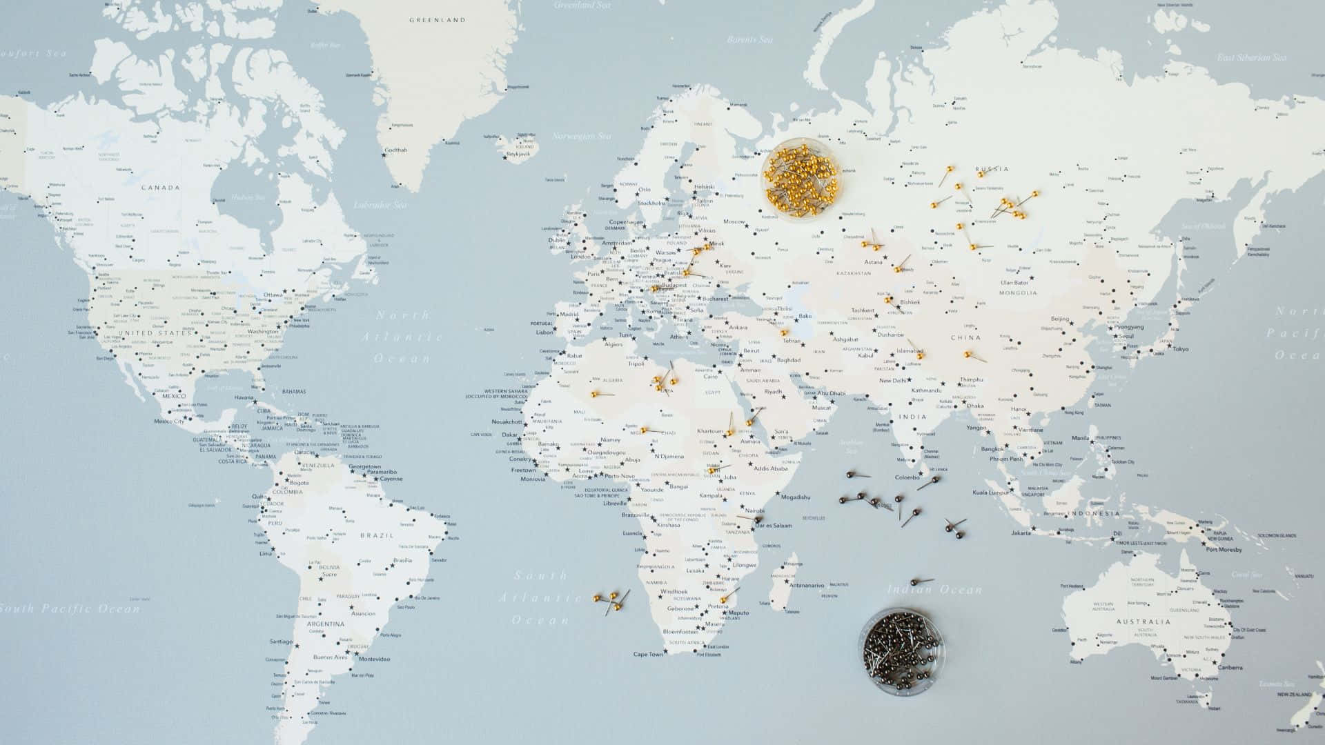 Map 桌布 壁紙 — 一個極簡風格的世界地圖，散落著金色和黑色的圖釘，在柔和的藍色背景上標記全球旅行目的地。