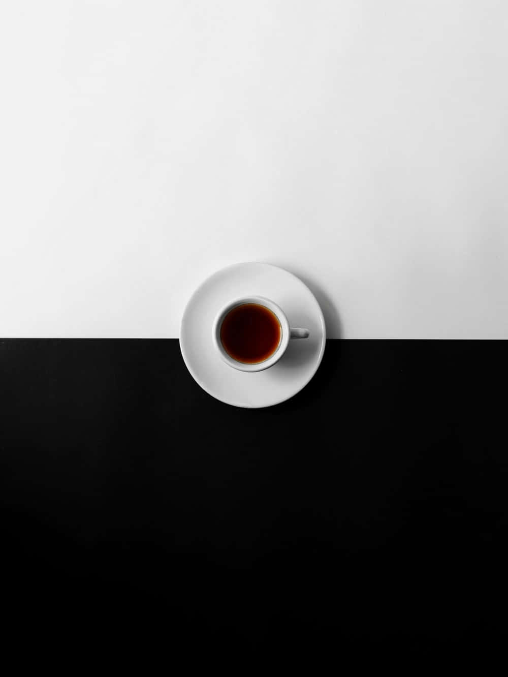 Minimalist Coffee 桌布 壁紙 — 一個極簡風格的俯拍鏡頭，展示一個白色茶杯裝著深色茶，放在碟子上，位於鮮明的黑白分割背景中央。