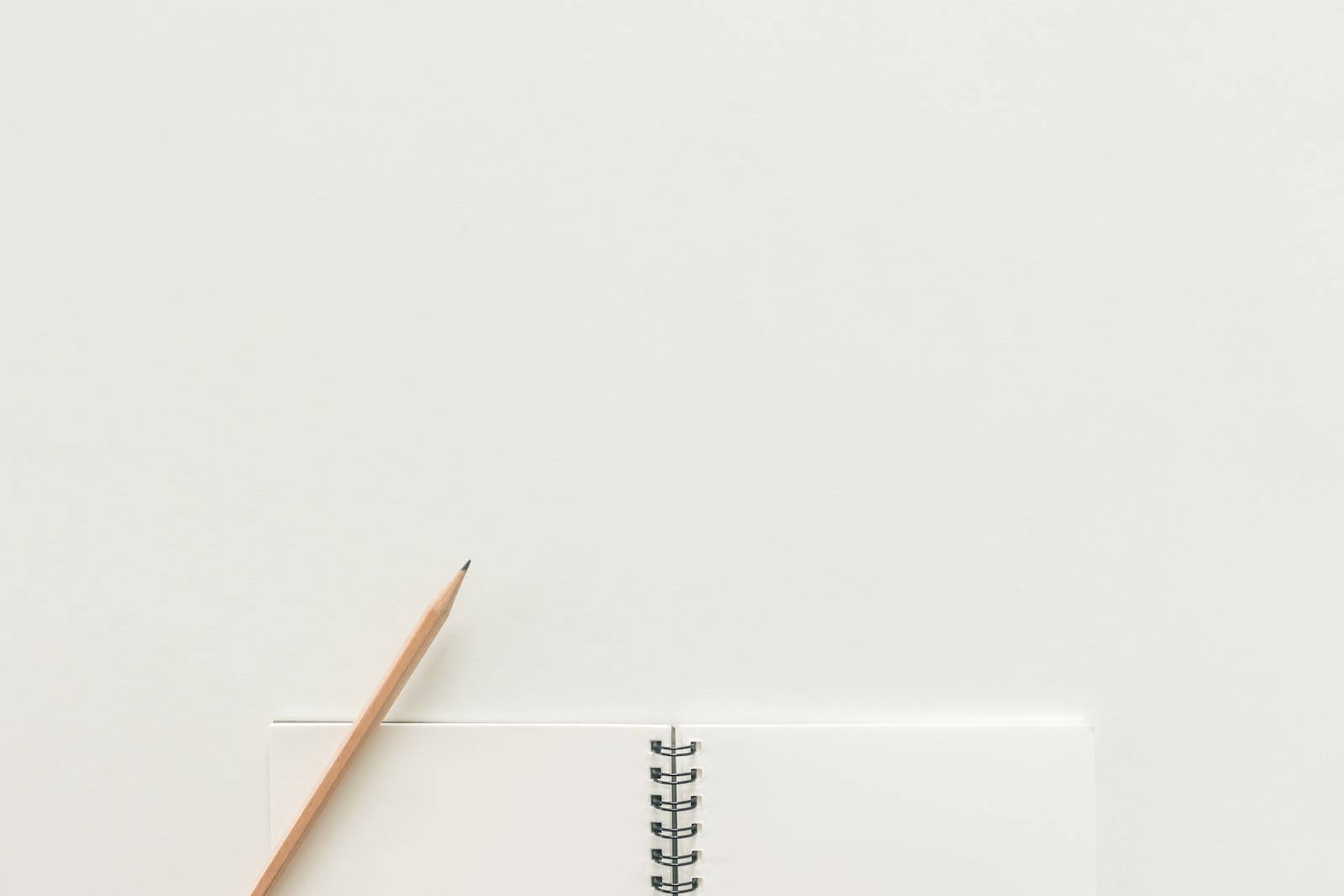 Notebook 桌布 壁紙 — 一個極簡風格的俯視圖，顯示一支木鉛筆放在一個打開的螺旋筆記本旁邊，背景是純白色表面。