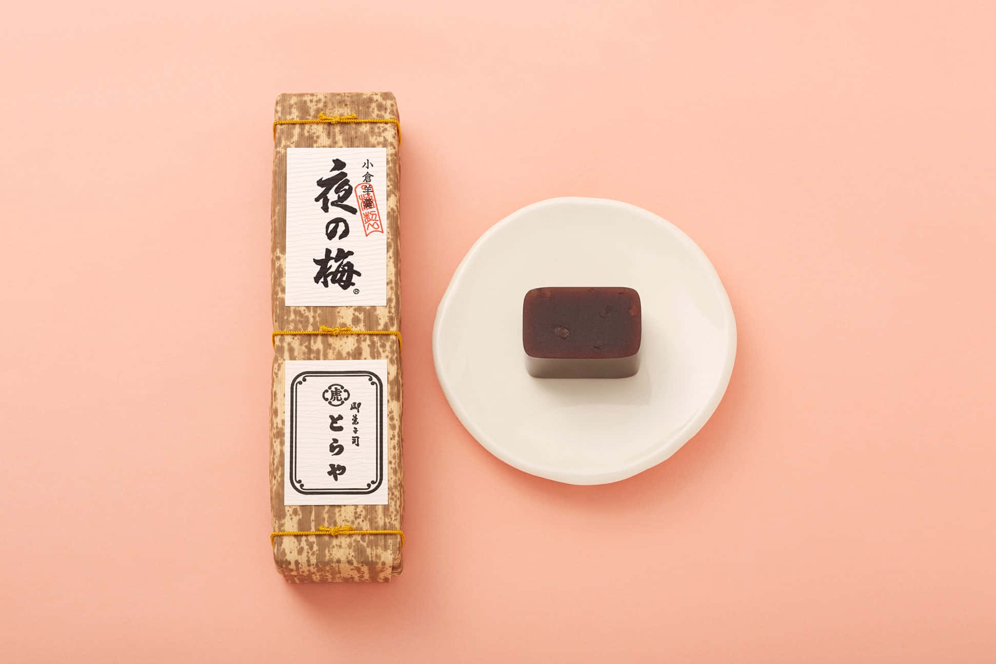 Japanese Sweets 桌布 壁紙 — 一個極簡風格的平鋪構圖，展示傳統日本梅子醬包裝和一個深棕色方形糖果放在白色盤子上，背景是柔和的粉紅色。