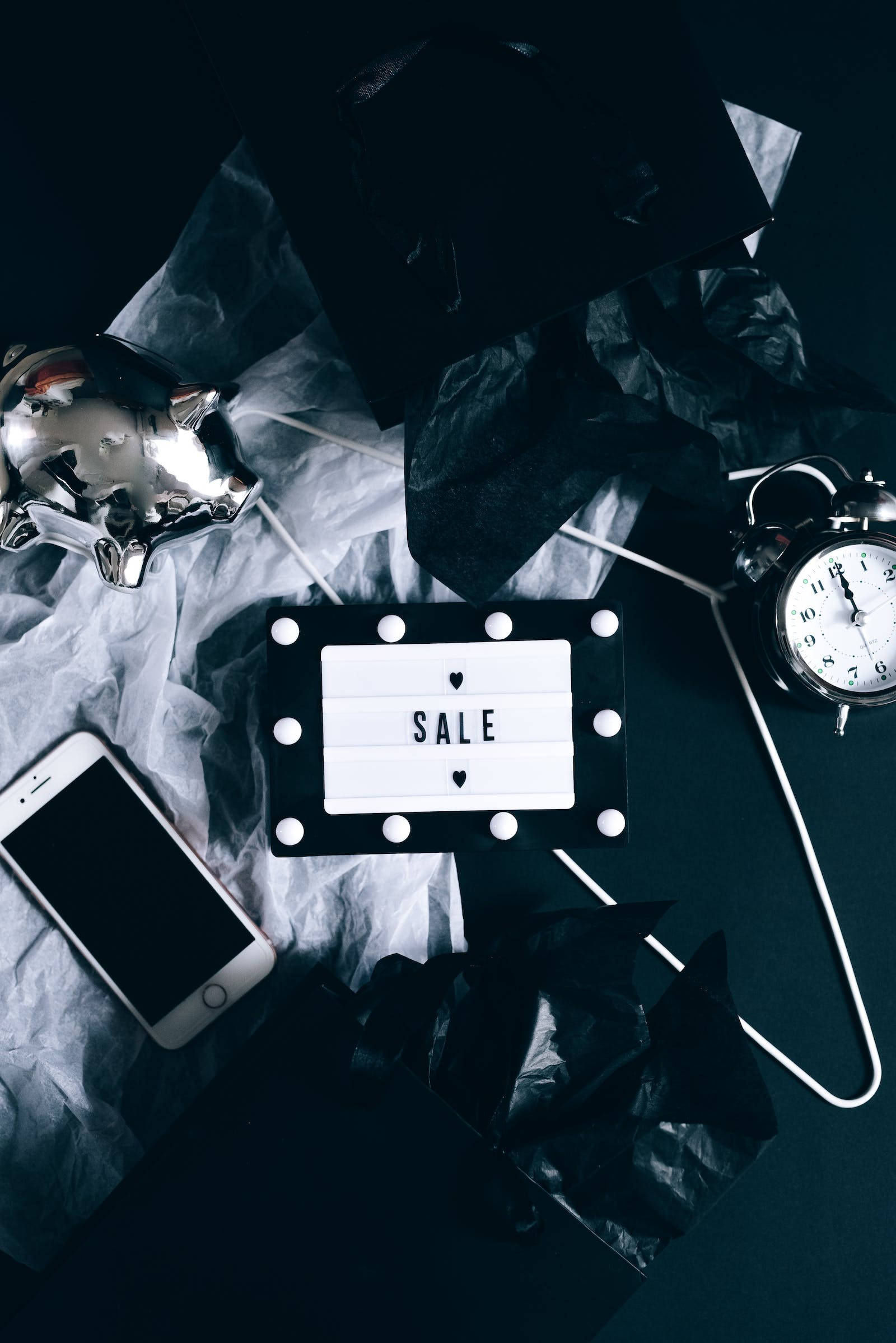 深色 iPhone 桌布 壁紙 — 一個極簡風格的平鋪構圖，發光的「SALE」標誌周圍環繞著皺巴巴的黑白紙張、一個復古鬧鐘和一部智慧型手機，置於深色表面上。