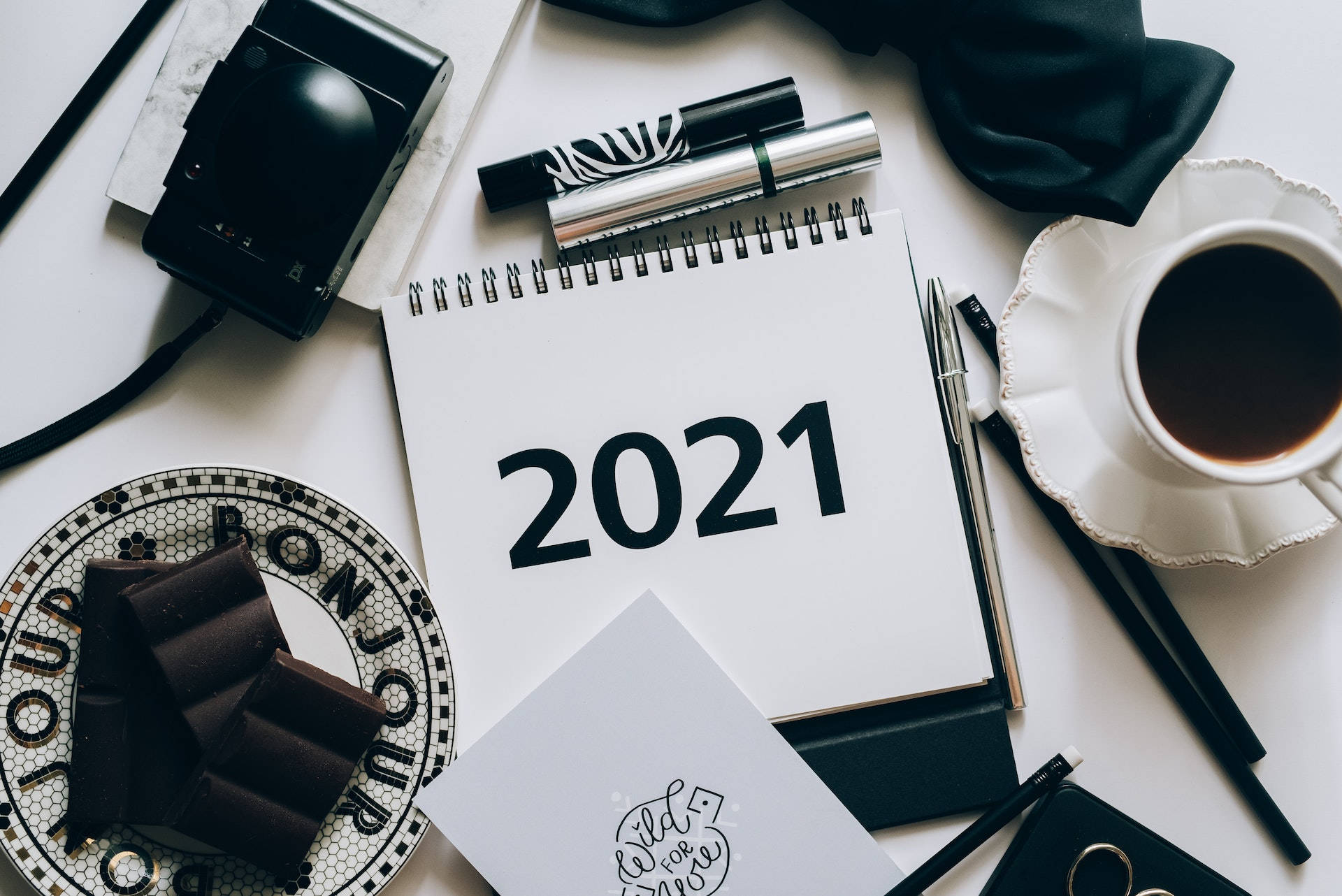 2021 桌面 桌布 壁紙 — 一個極簡風格的平面佈置，包含一本2021年的筆記本，周圍有咖啡、巧克力、筆和一部復古相機，放在白色表面上。