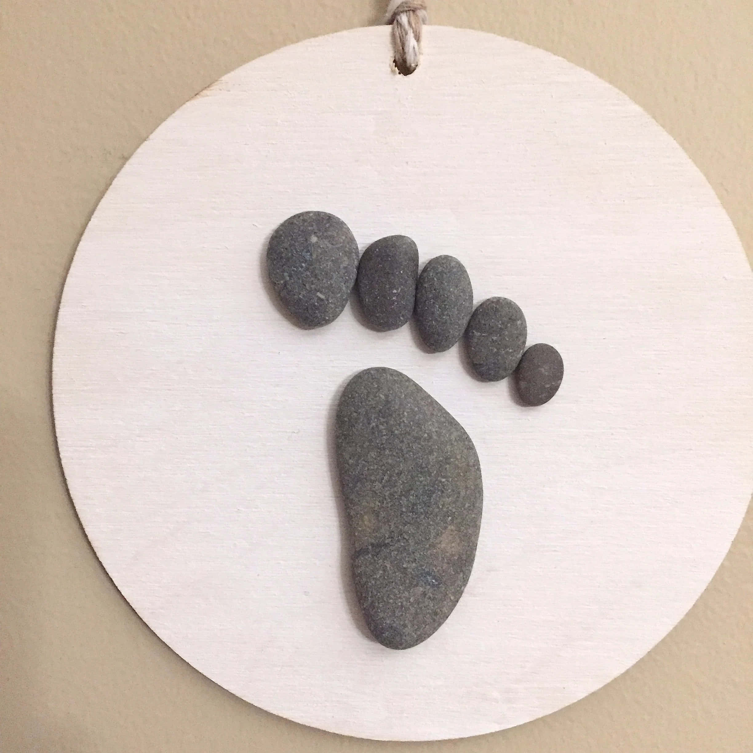 Pebble Art 桌布 壁紙 — 一個極簡風格的牆壁掛飾，由光滑的灰色河石排列在圓形白色木盤上形成腳印圖案。