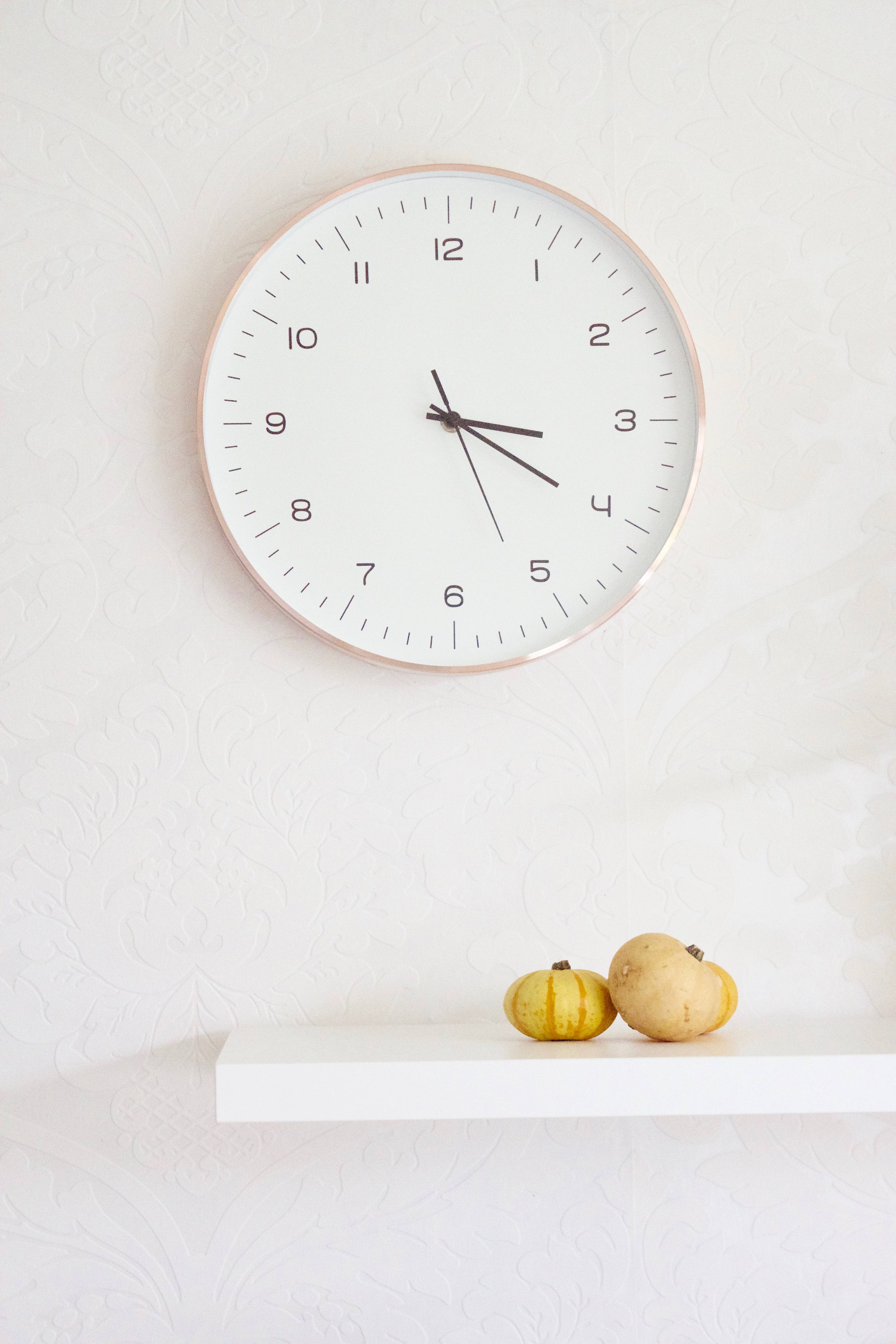 Clock 桌布 壁紙 — 一個極簡風格的白色掛鐘，帶有玫瑰金邊框，掛在一個放有兩個小南瓜的架子上方。