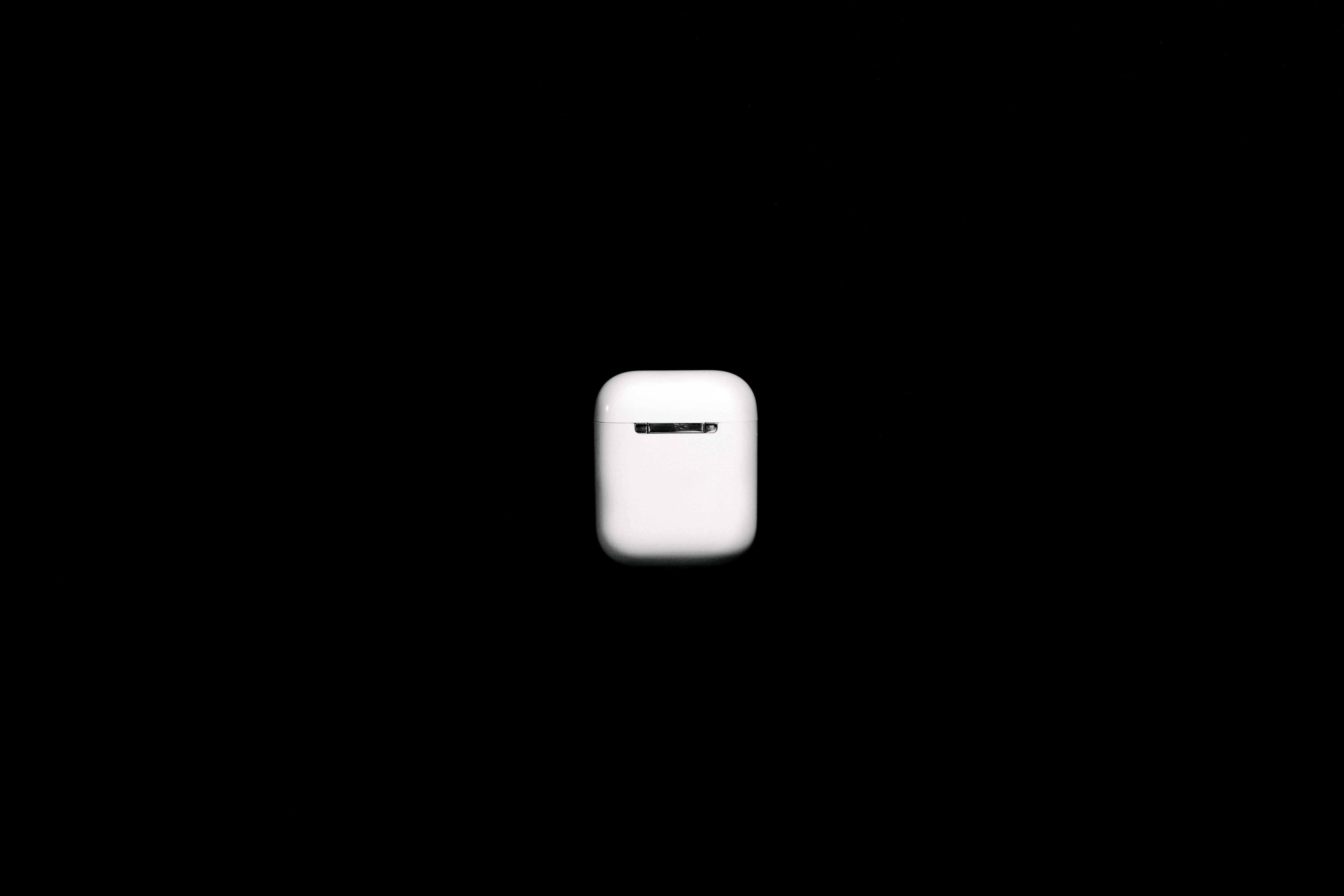 AirPods 桌布 壁紙 — 一個極簡風格的白色無線耳機充電盒，置於純黑色背景中央，帶有柔和燈光。