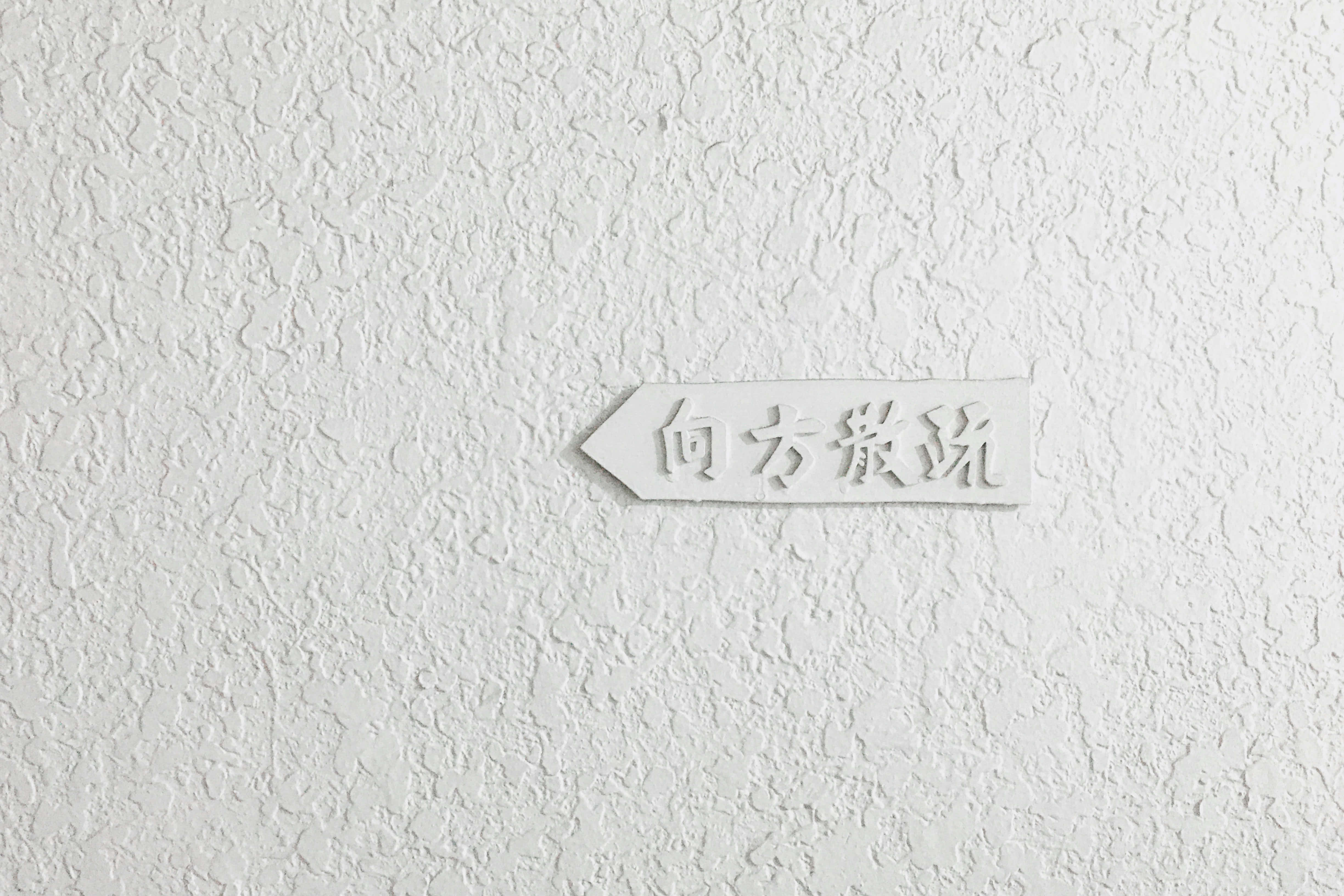 White Texture 桌布 壁紙 — 一個極簡風格的白色箭頭標誌，上面有日文字元，安裝在帶有紋理的灰泥牆上。