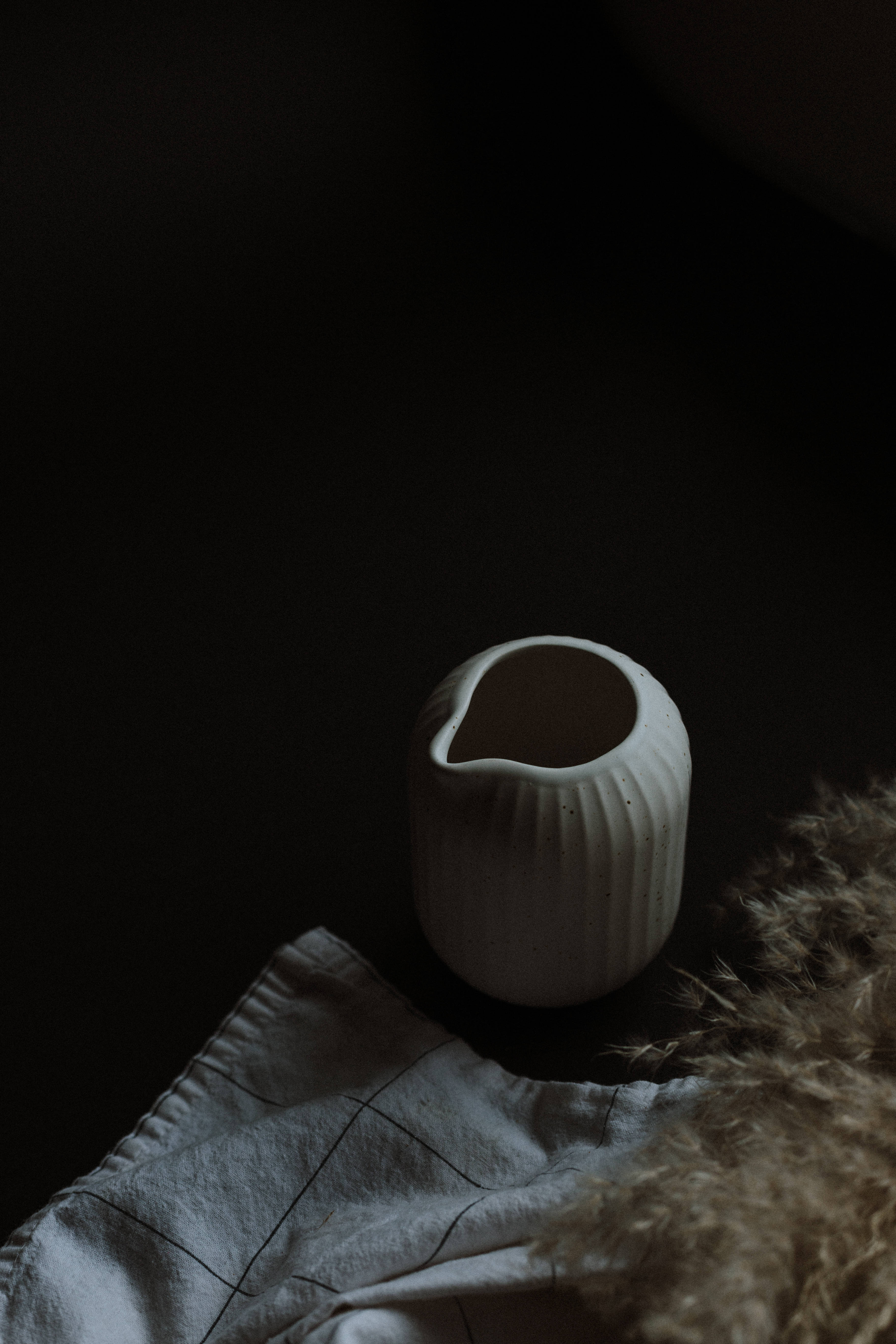 simple dark aesthetic 桌布 壁紙 — 一個極簡風格的白色陶瓷壺，帶有垂直條紋，放在格紋布上，旁邊是蓬鬆的棕色毛皮，背景為深色。