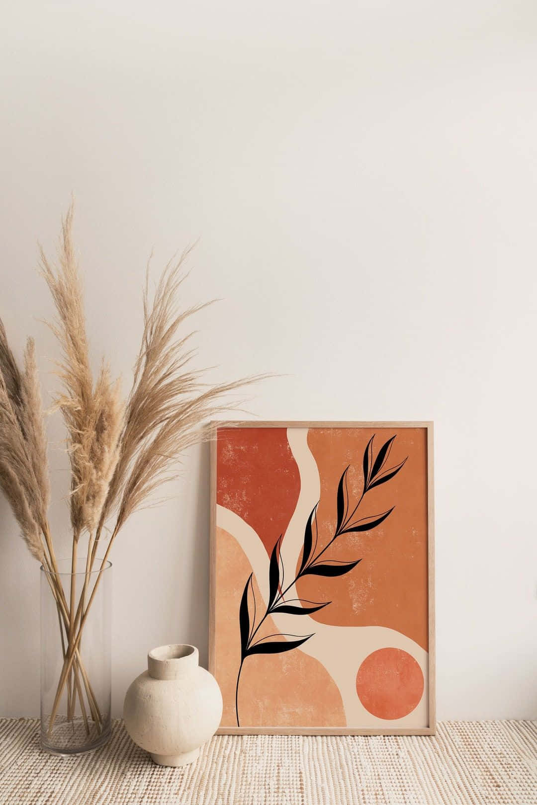 Minimalist Painting 桌布 壁紙 — 一個極簡風格的裱框抽象藝術品，帶有陶土色和奶油色形狀以及黑色葉子剪影，旁邊放著乾燥的蒲葦和一個陶瓷花瓶，置於編織地毯上。
