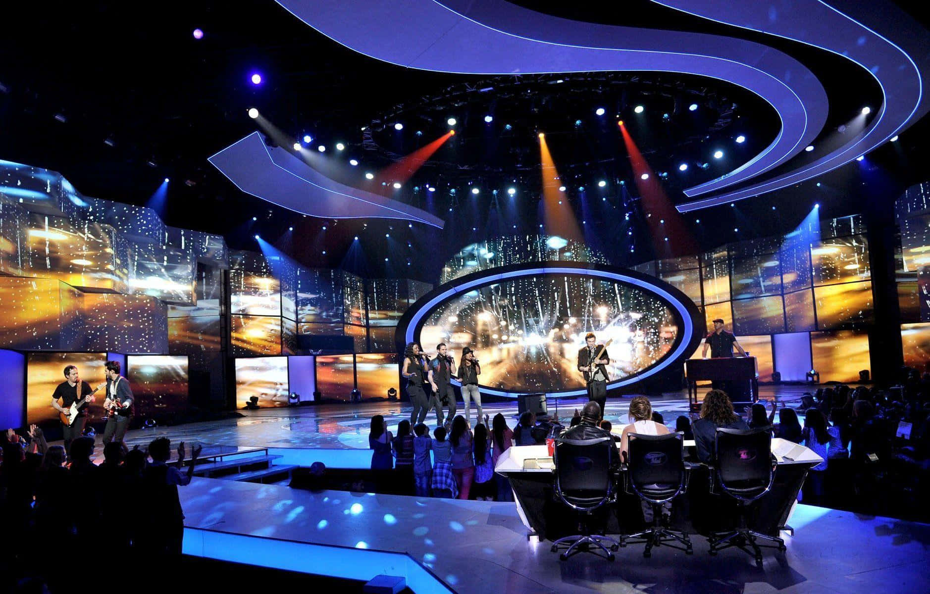 american idol 桌布 壁紙 — 一個樂團在未來風格的舞台上表演，發光的藍色曲線和溫暖的橙色聚光燈照亮了音樂家和評審。