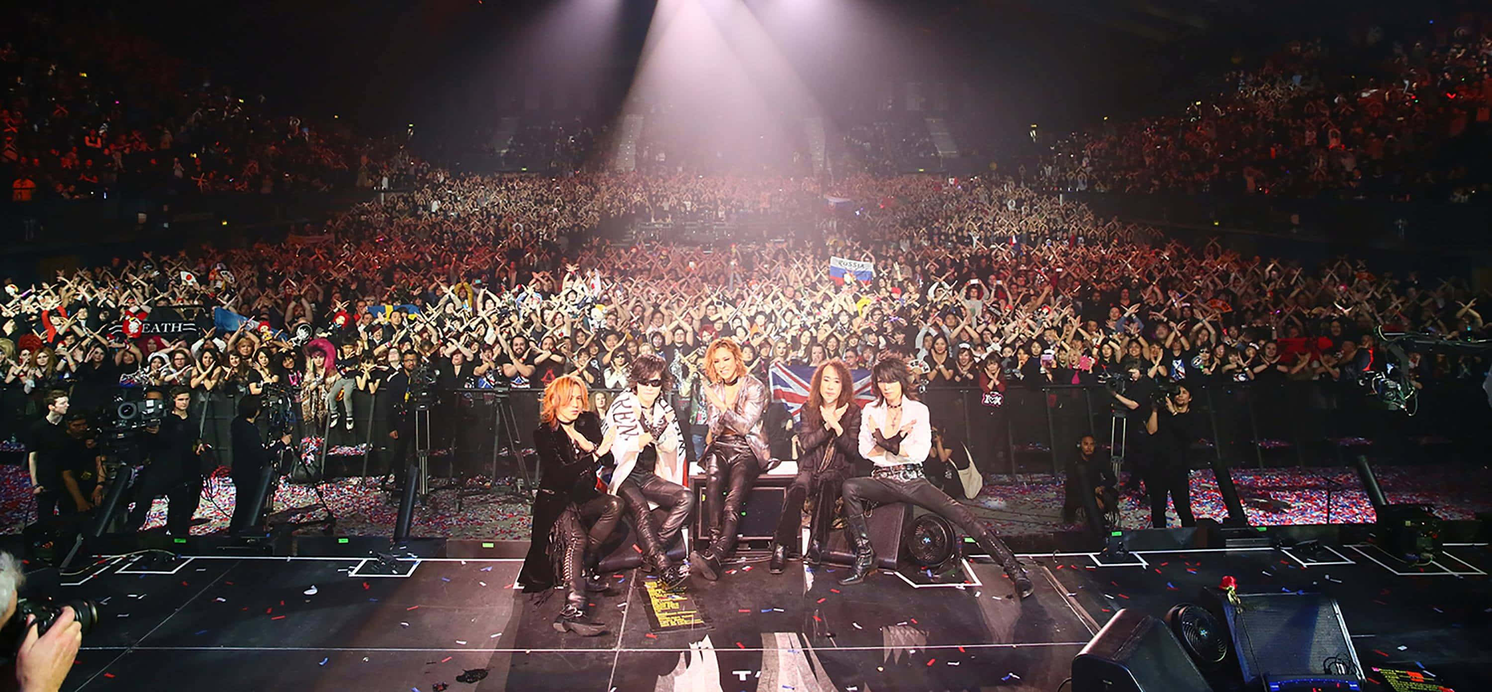 Visual Kei 桌布 壁紙 — 一個樂團在燈光璀璨的舞台上表演，面對著擠滿場館、揮舞著螢光棒的龐大群眾。