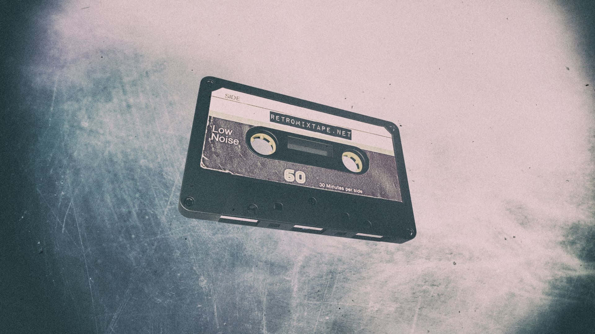 Mixtape 桌布 壁紙 — 一個標有「RETROMIXTAPE.NET」的復古黑色卡式錄音帶，放在一個有刮痕的金屬表面上，背景是褪色的藍白漸變色。