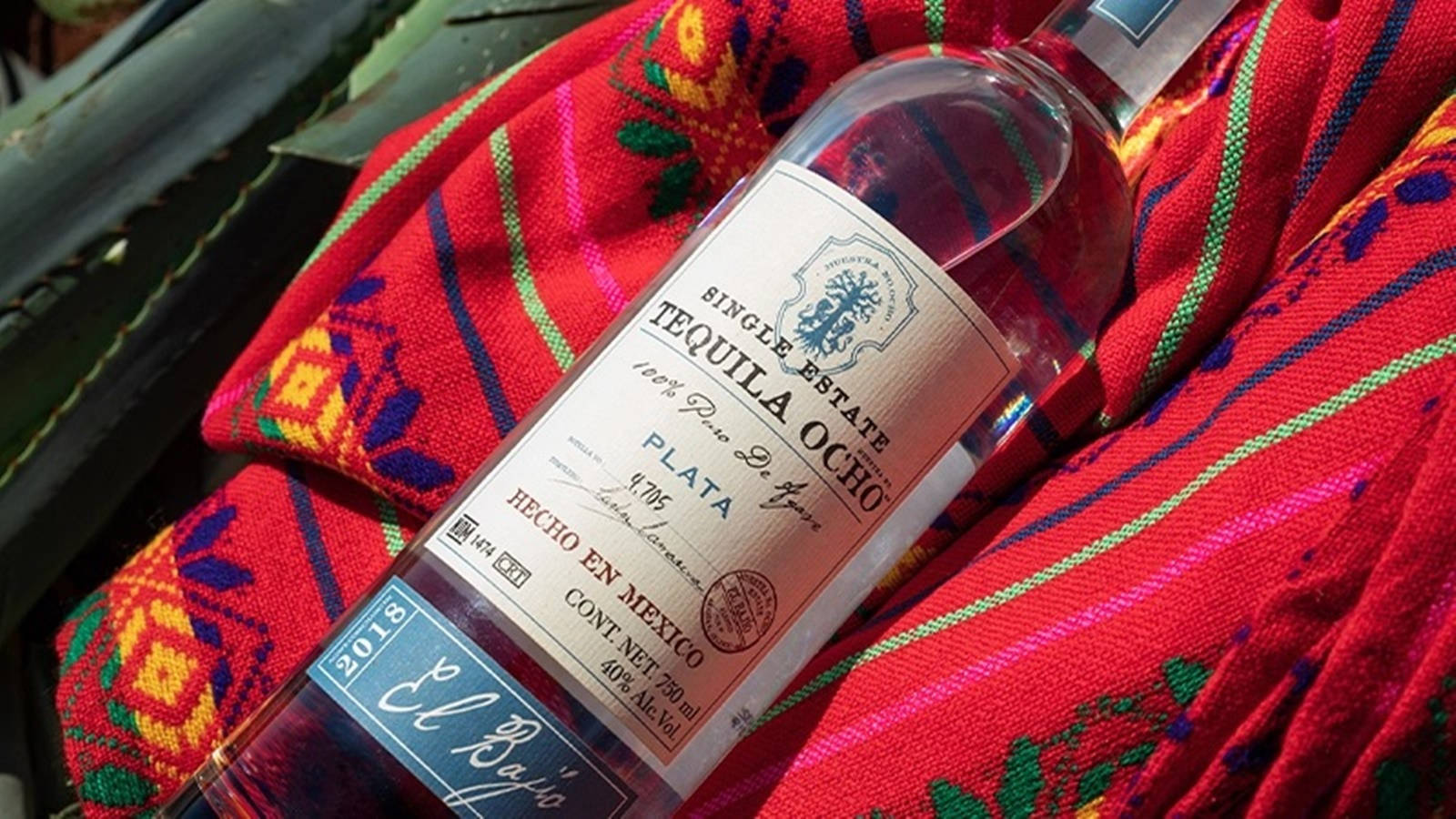 bottle 桌布 壁紙 — 一個標有「Single Estate Tequila Ocho」的透明龍舌蘭酒瓶，斜放在一塊帶有綠色和黃色圖案的鮮紅色刺繡布料上。
