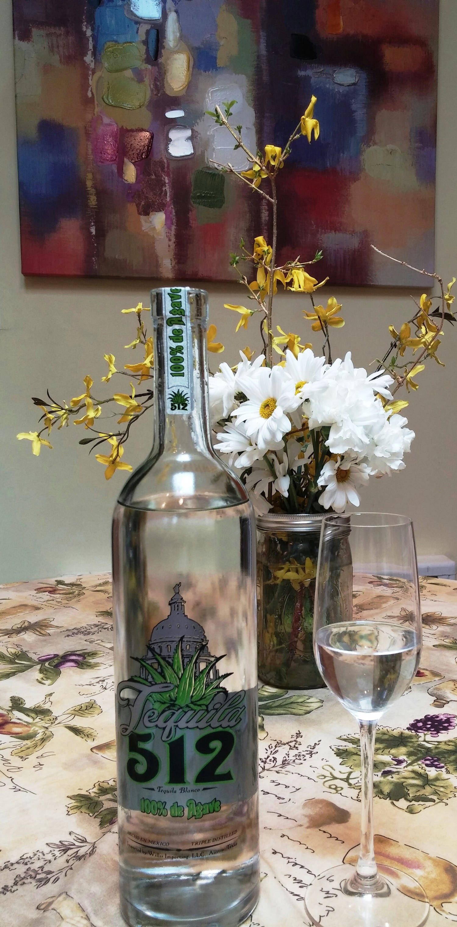 drinks 桌布 壁紙 — 一個標有512的透明龍舌蘭酒瓶，旁邊放著一個酒杯和一個花瓶，裡面插著白色雛菊和黃色連翹，放在花卉桌布上。