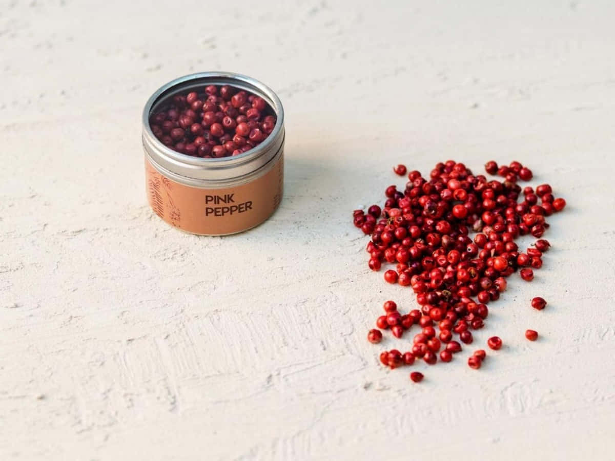 Pink Peppercorn 桌布 壁紙 — 一個標有粉紅胡椒的小罐子，將鮮紅的胡椒粒灑在帶有紋理的白色表面上。