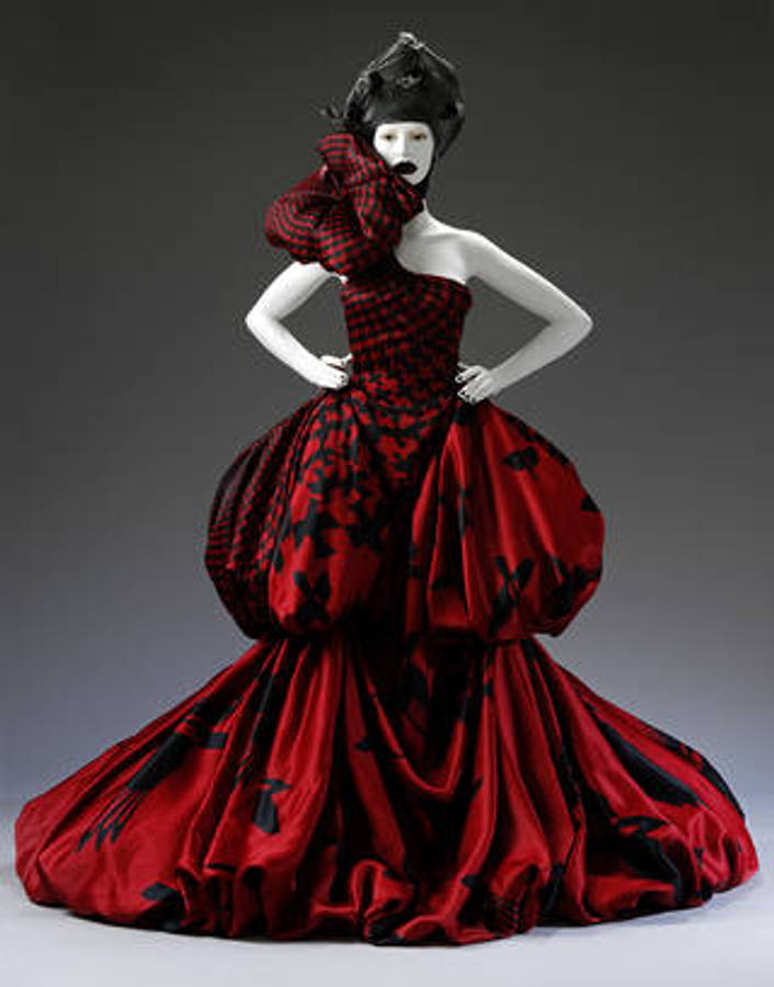Alexander Mcqueen 桌布 壁紙 — 一個模特兒穿著一件戲劇性的紅黑格紋長裙，搭配寬大的裙子和相配的帽子，背景是深灰色的攝影棚。
