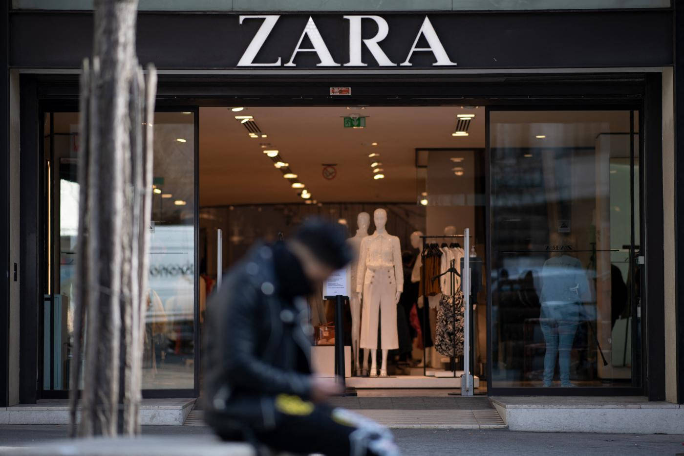 Zara 桌布 壁紙 — 一個模糊的人穿著黑色外套坐在Zara商店外面，玻璃門內可見人體模型和衣服架。