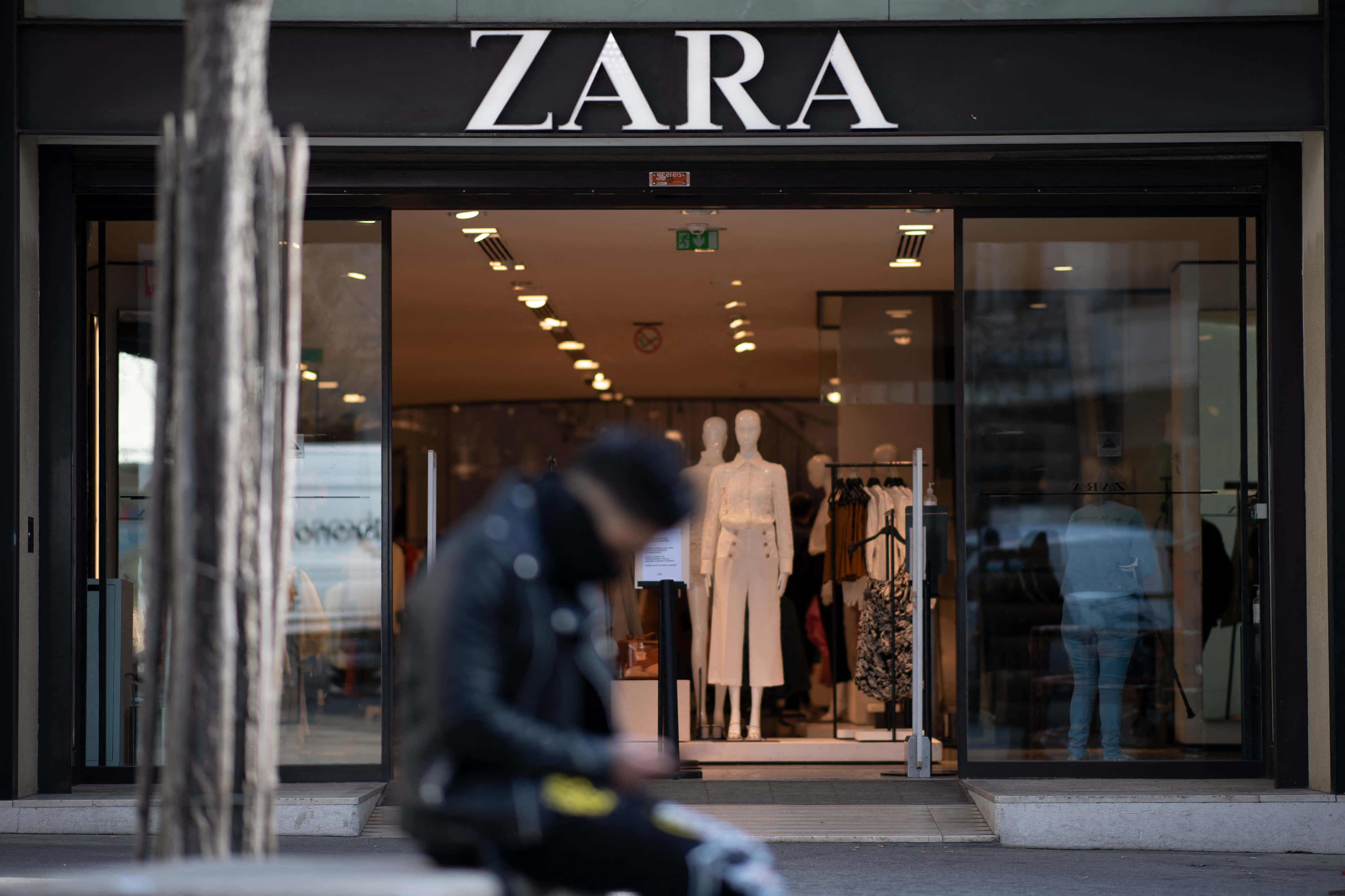 Zara 桌布 壁紙 — 一個模糊的人穿著黑色外套坐在Zara商店外面，裡面有模特兒展示白色服裝。