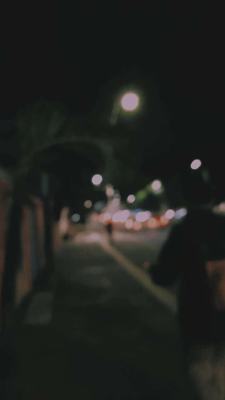 城市 桌布 壁紙 — 一個模糊的夜景，一個人走在昏暗的城市人行道上，有散景路燈和遠處的交通。