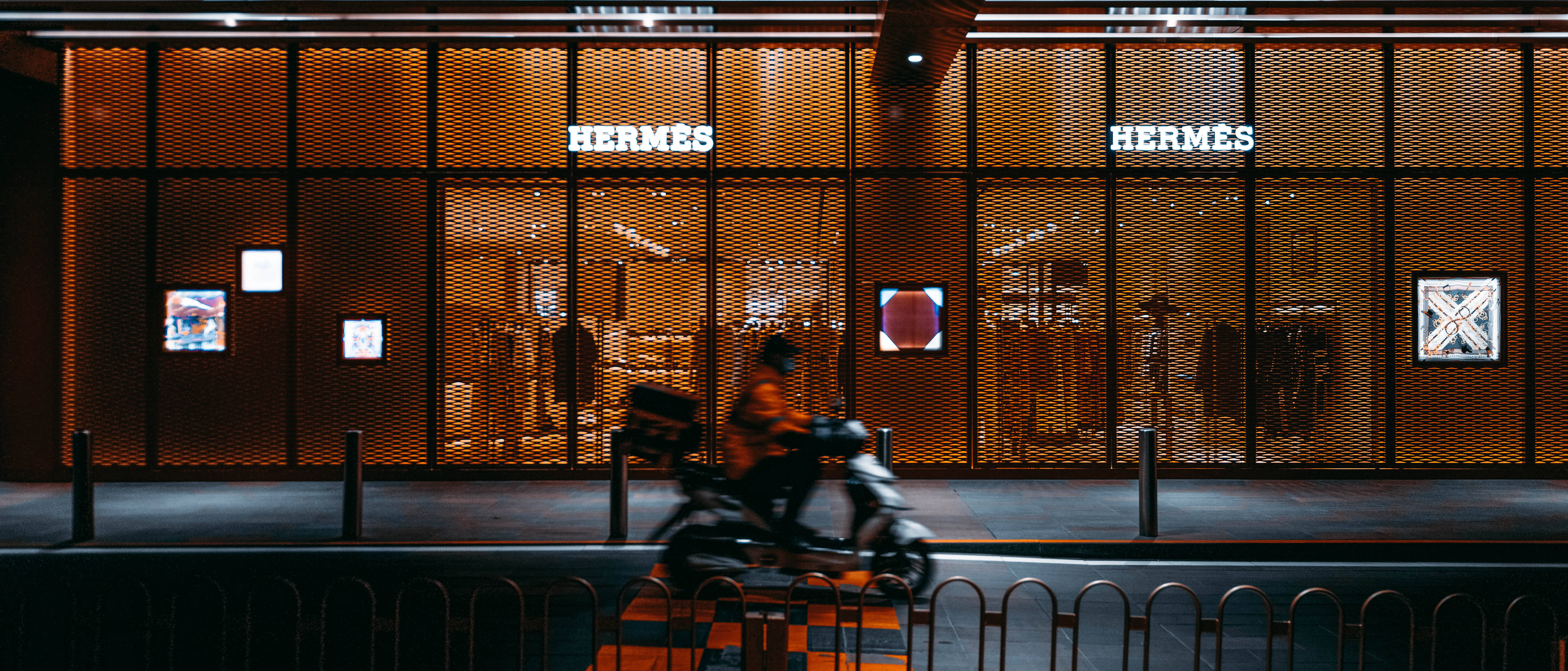 Hermes 桌布 壁紙 — 一個模糊的機車騎士在夜間滑過一間溫暖照明的愛馬仕商店，帶有發光的招牌和圖案外牆。