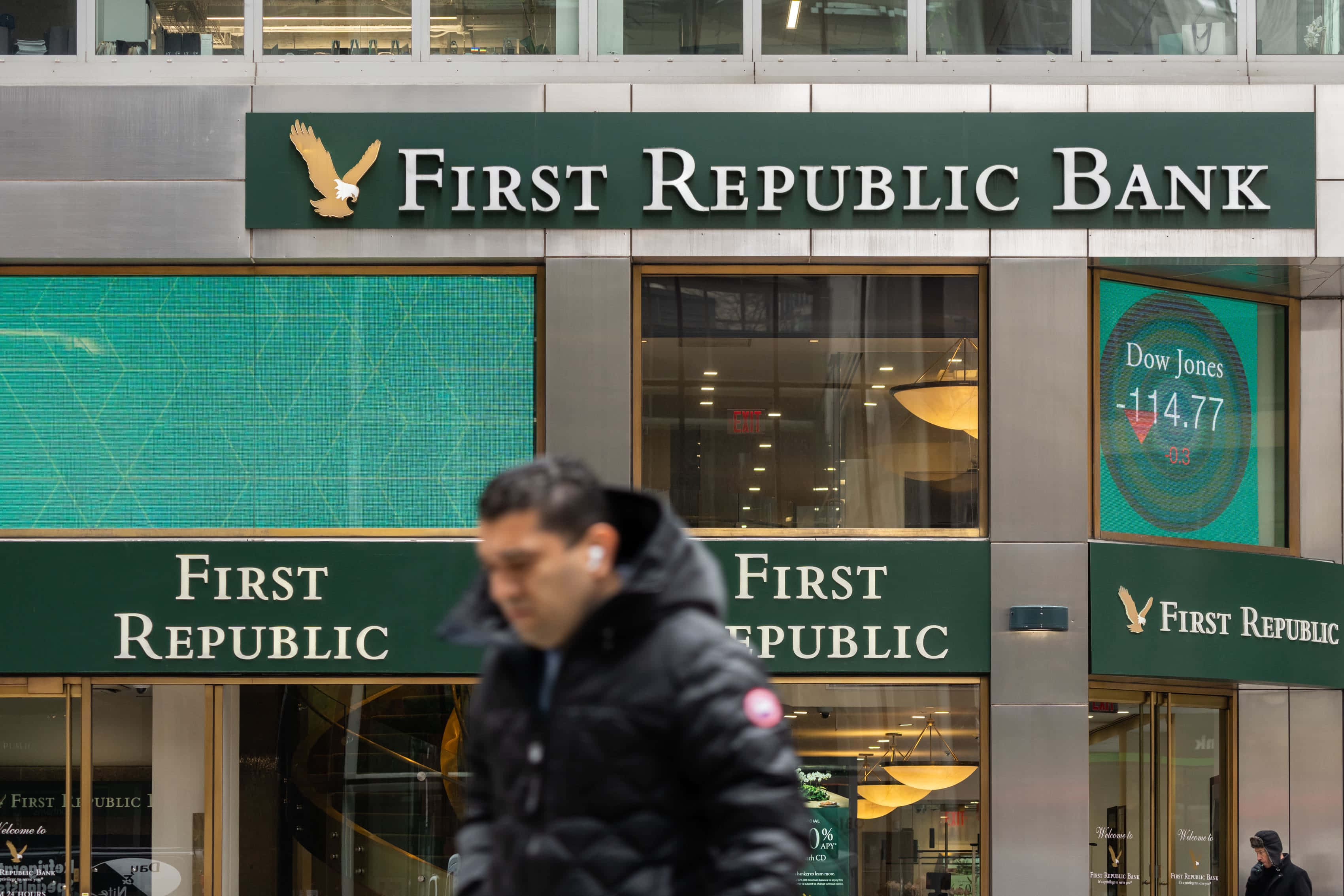 Bank 桌布 壁紙 — 一個模糊的行人走過第一共和銀行大樓，其數位螢幕顯示道瓊斯指數下跌114.77點。
