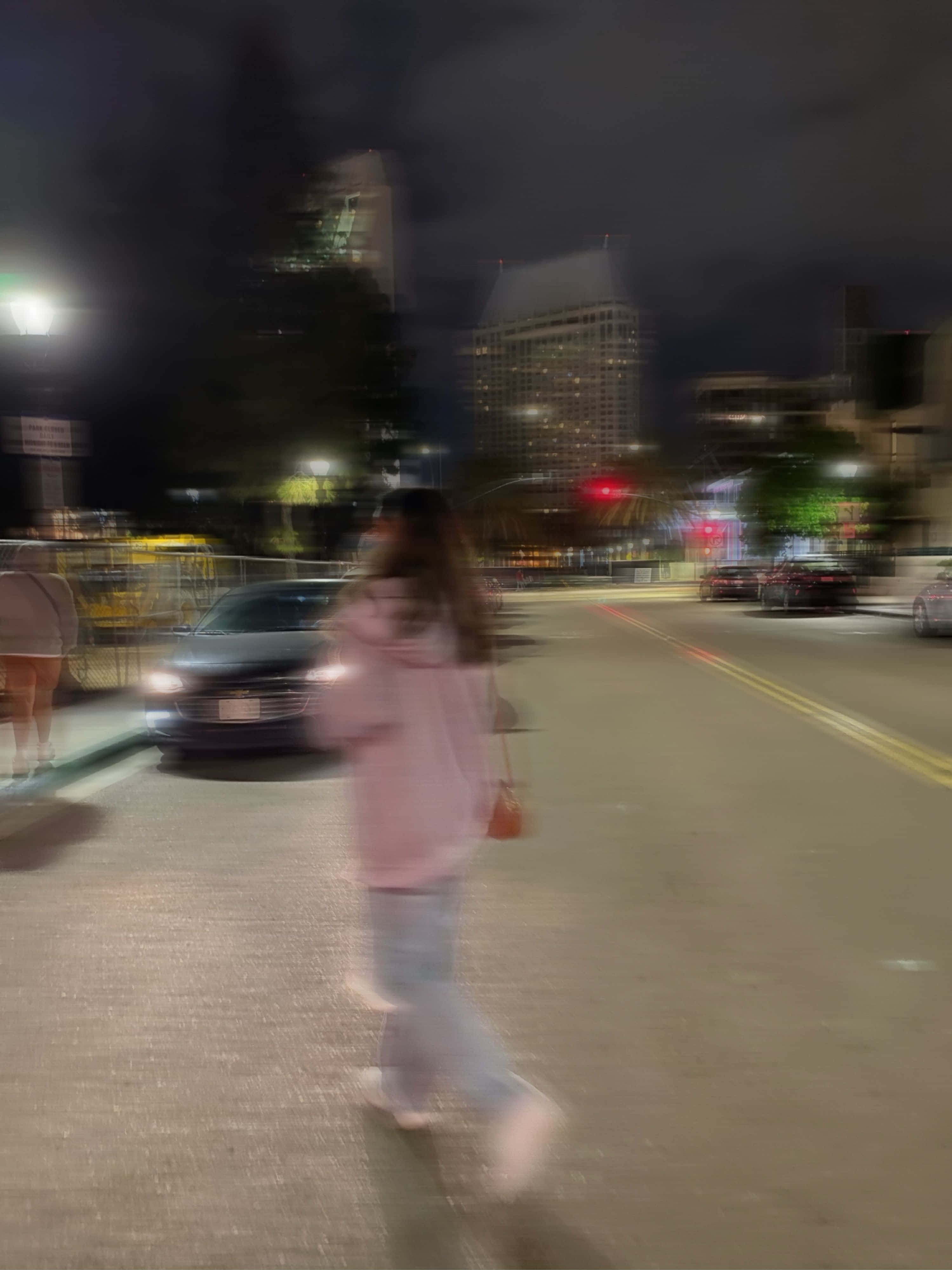 女孩 桌布 壁紙 — 一個模糊的身影穿著粉紅色連帽衫，在夜晚的城市街道上行走，背景有發光的車頭燈和高樓大廈。