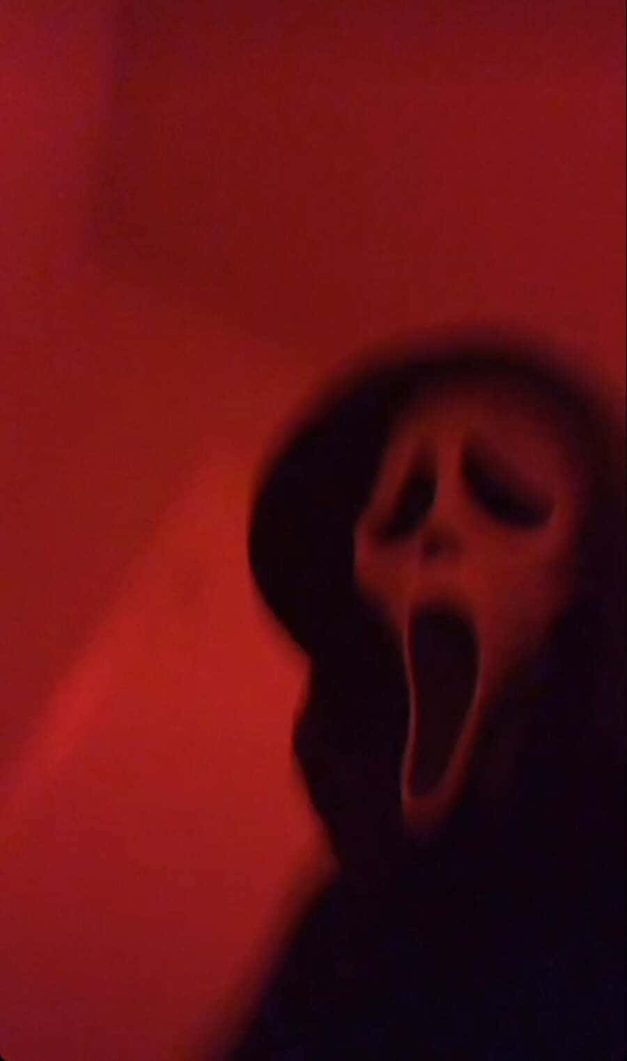 Ghostface Aesthetic 桌布 壁紙 — 一個模糊的鬼影戴著白色尖叫面具，在陰暗的紅光中站在黑暗的牆壁前。