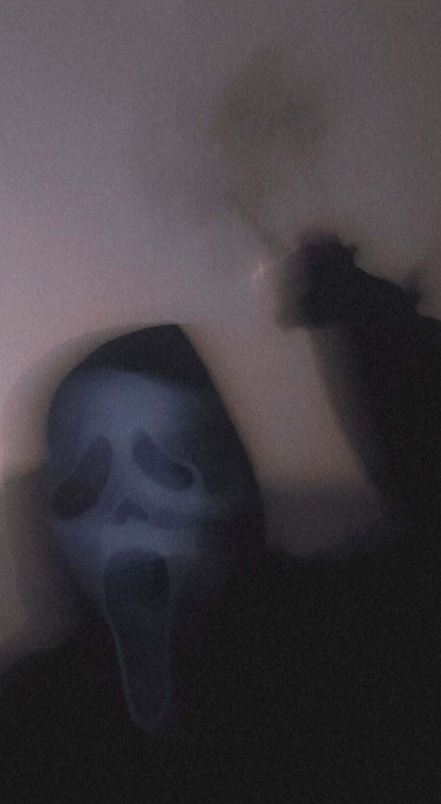 Ghostface Aesthetic 桌布 壁紙 — 一個模糊的鬼面具，眼睛睜大，嘴巴張開，在黑暗的房間裡發出微弱的光芒。