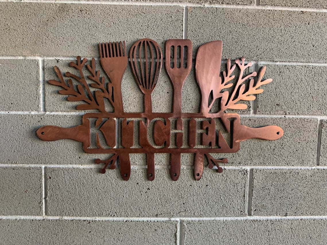 Kitchen Wall 桌布 壁紙 — 一個樸實的銅製廚房標誌，配有餐具和葉狀裝飾，安裝在灰色磚牆上。