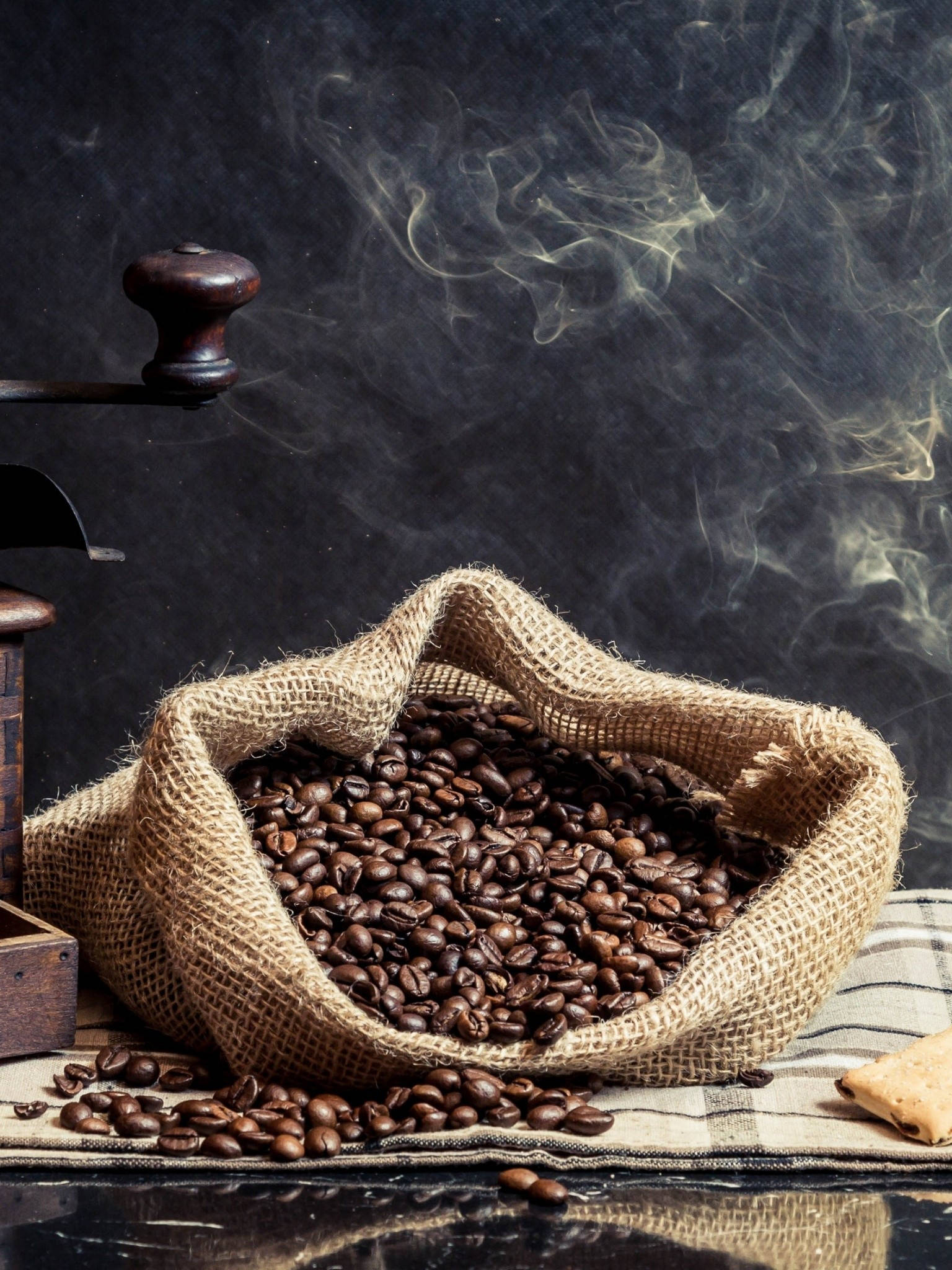 beans 桌布 壁紙 — 一個樸素的麻布袋裝滿了烘焙咖啡豆，旁邊放著一個木製研磨機，香氣四溢的蒸汽在黑暗背景下裊裊升起。