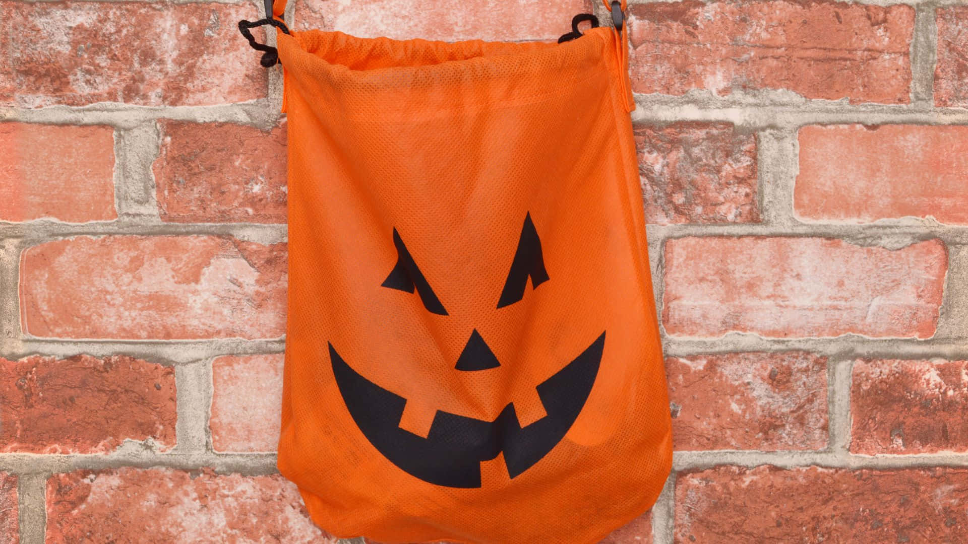 Trick Or Treat Bags 桌布 壁紙 — 一個橘色的萬聖節trick-or-treat袋，上面有黑色的jack-o'-lantern臉，掛在紅磚牆上。
