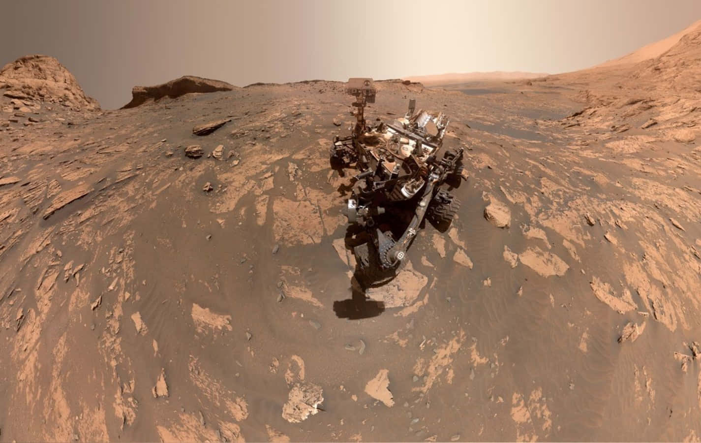 Mars Surface 桌布 壁紙 — 一個機器人探測車站在火星鏽紅色的表面上，在朦朧的天空下將陰影投射在岩石地形上。