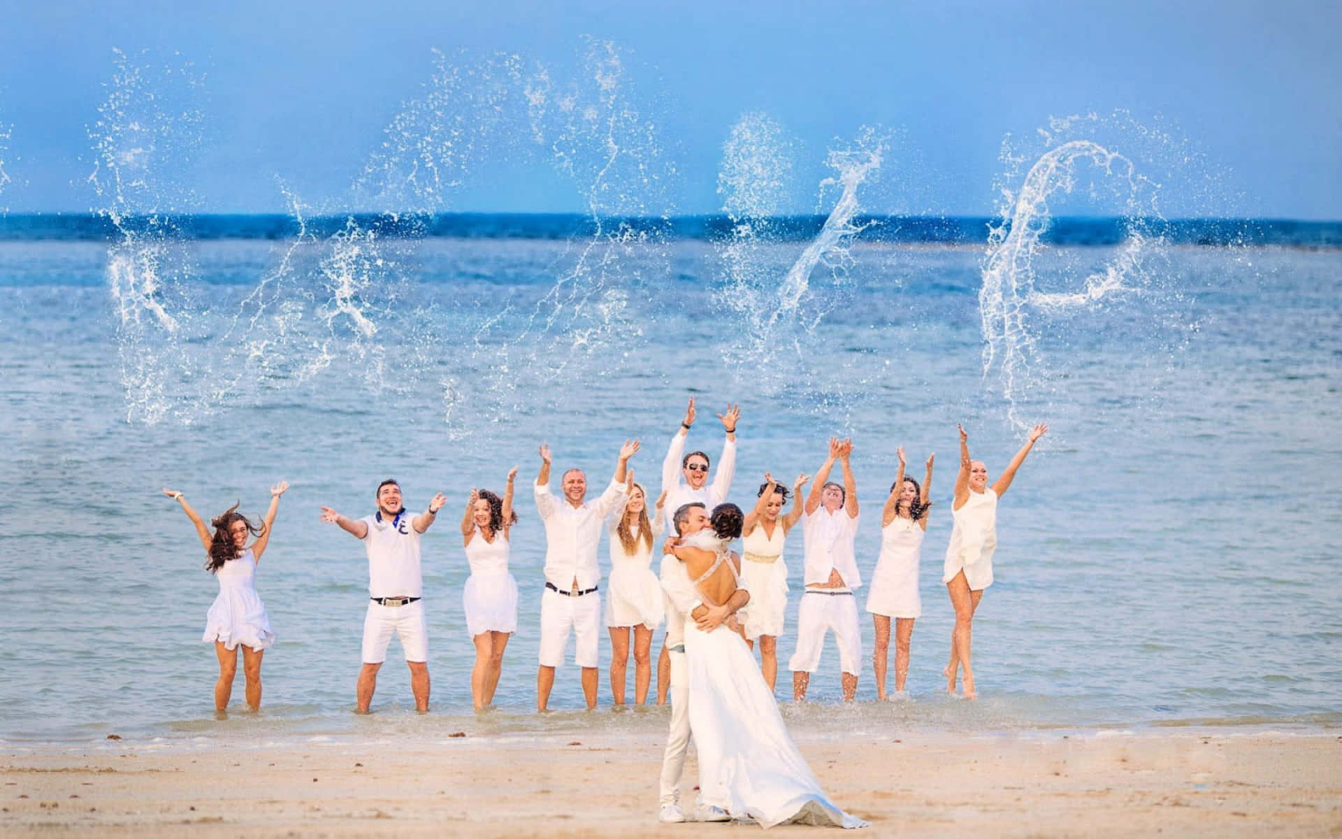 Beach Scenes Desktop 桌布 壁紙 — 一個歡樂的婚禮派對穿著白色服裝站在陽光明媚的海灘上，水花飛濺在他們上方形成love這個詞。