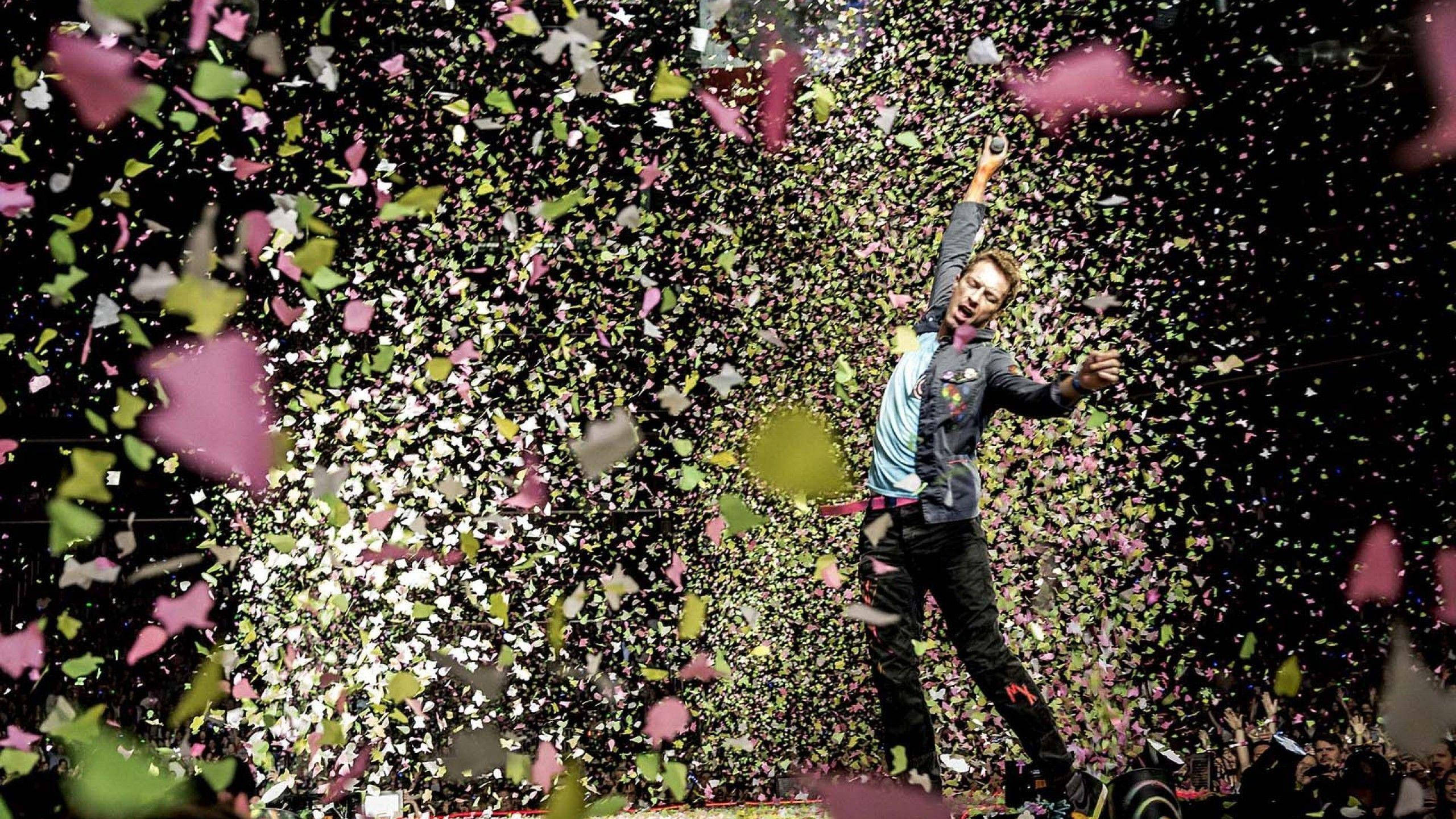 Chris Martin 桌布 壁紙 — 一個歡欣鼓舞的男人在深色背景下慶祝，粉紅、綠色和白色的五彩紙屑風暴在他周圍爆發。