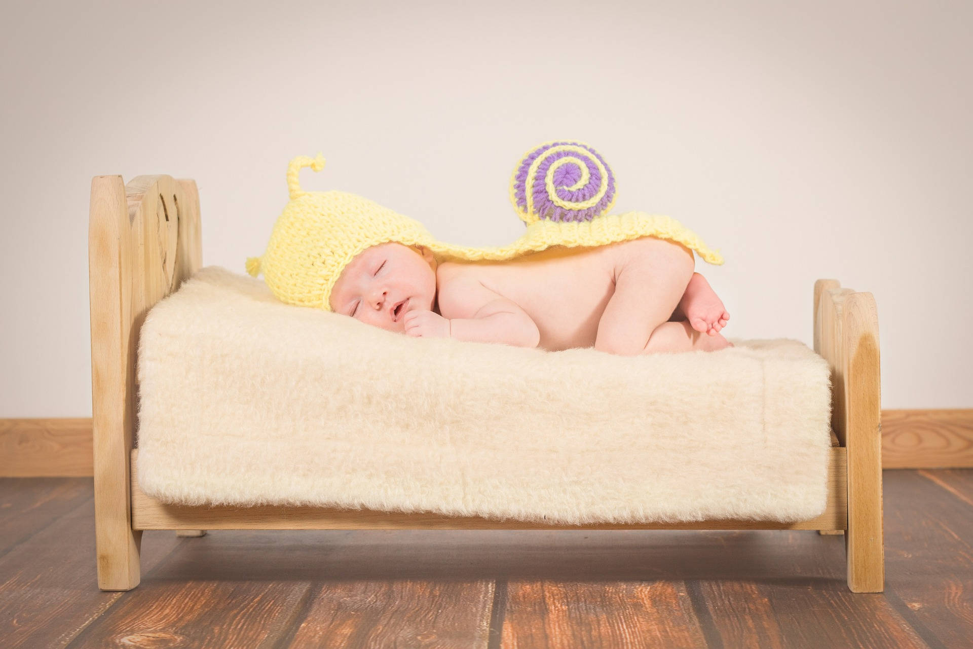 Very Cute Baby 桌布 壁紙 — 一個正在睡覺的新生嬰兒戴著黃色鉤針蝸牛帽，躺在鋪有柔軟奶油色毯子的木床上。