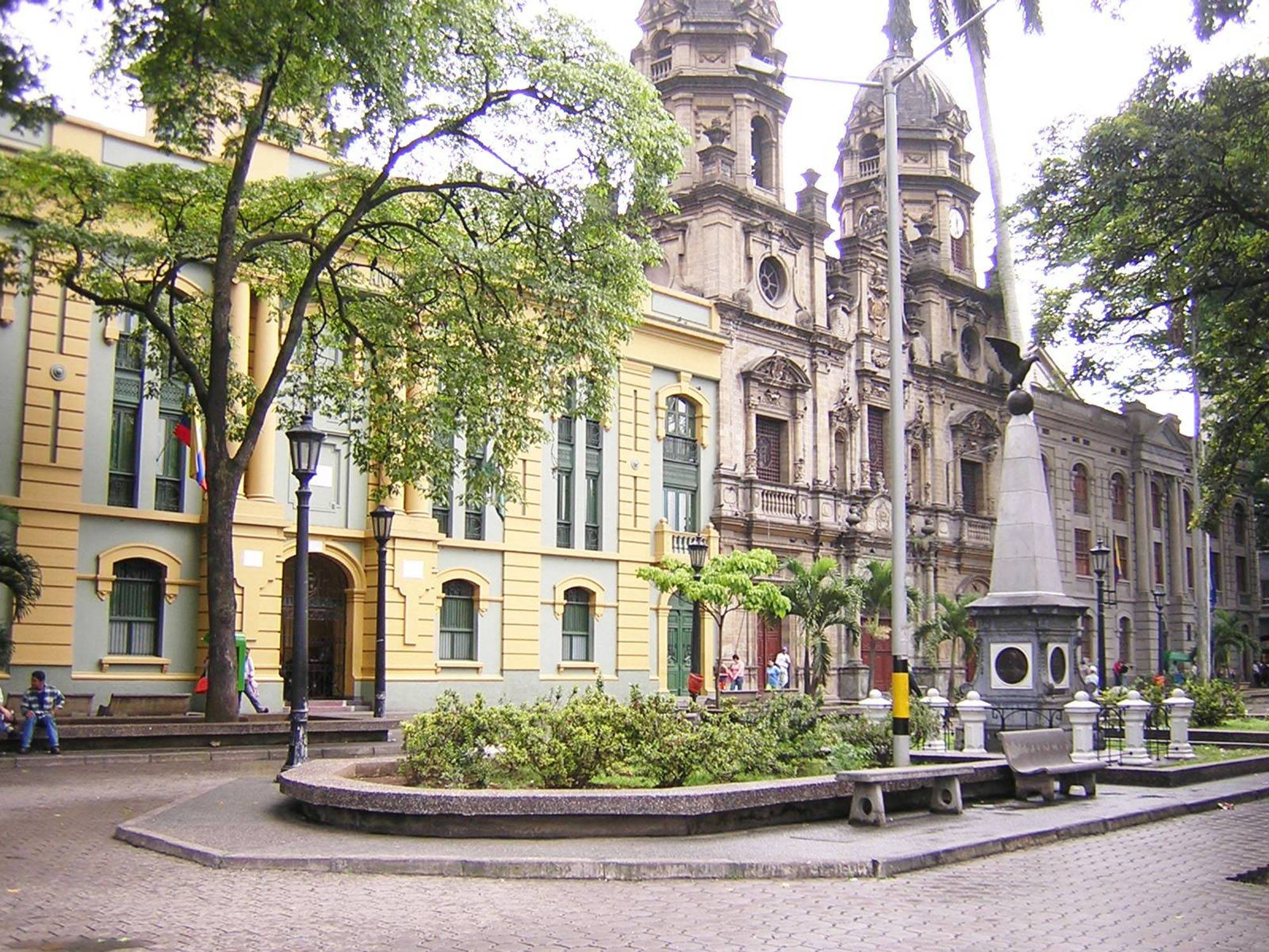 Medellin 桌布 壁紙 — 一個歷史悠久的廣場，擁有華麗的石建築、綠樹和一個中央紀念碑，周圍有長椅和行人。
