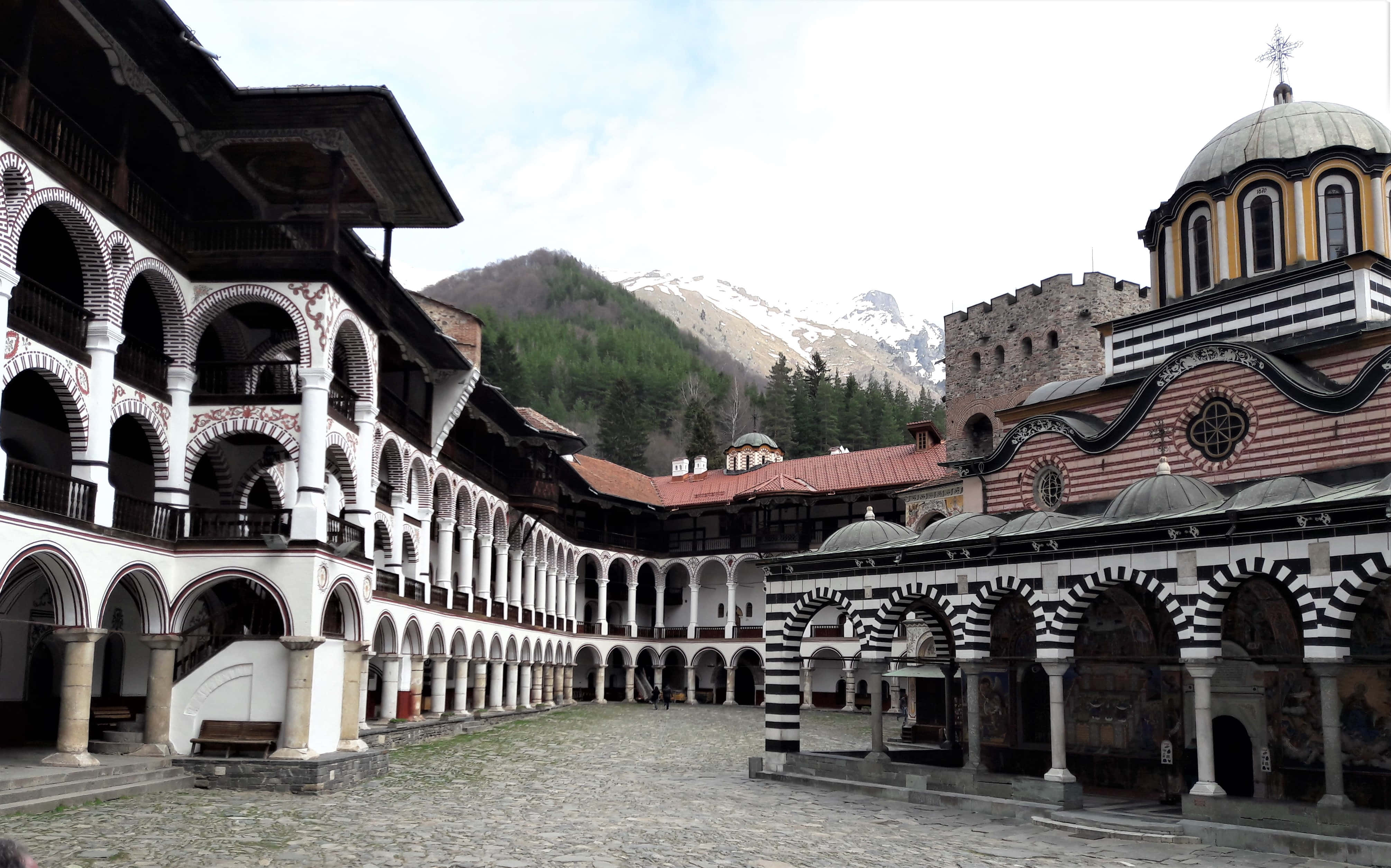 Bulgaria 桌布 壁紙 — 一個歷史悠久的東正教修道院的宏偉庭院，設有拱形柱廊，背後聳立著一座白雪覆蓋的山峰。