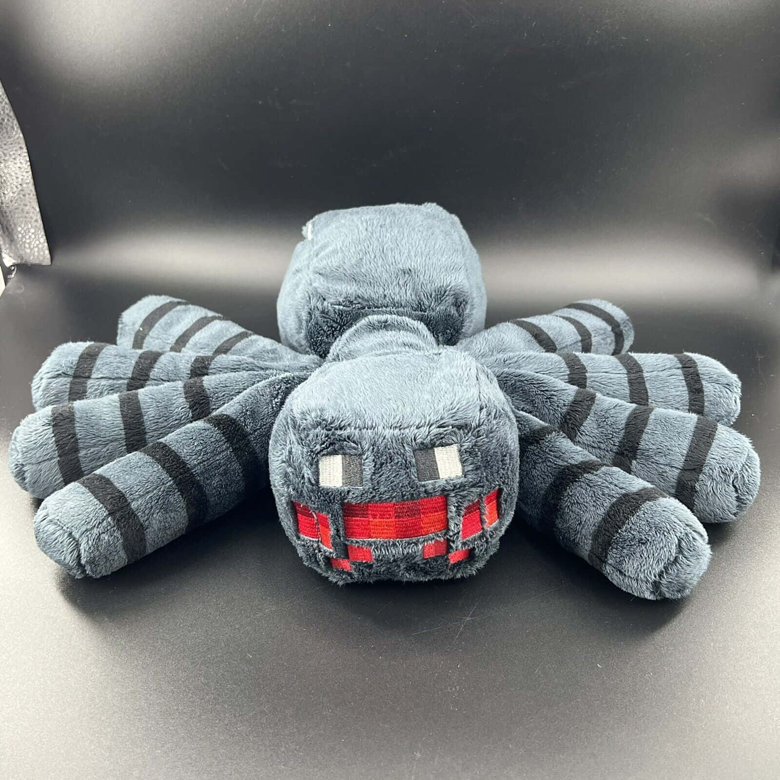 Minecraft Spider 桌布 壁紙 — 一個毛絨Minecraft蜘蛛玩具，有灰色和黑色條紋的腿和紅色嘴巴，坐在深灰色表面上。