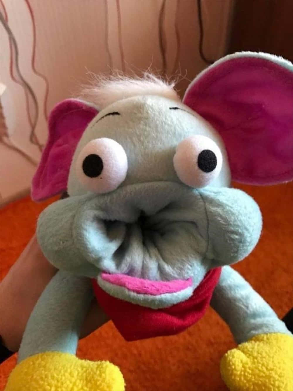 stuffed animal 桌布 壁紙 — 一個毛絨大象玩具的特寫，有粉紅色耳朵和黃色腳，被握在橙色地毯地板上。