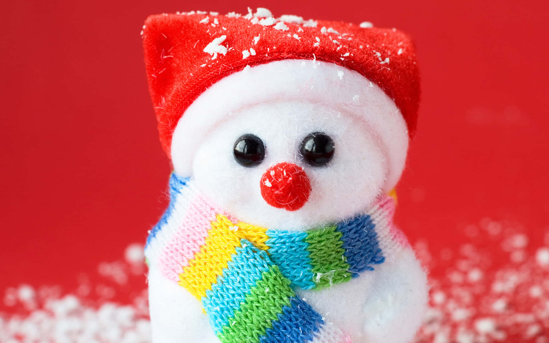  壁紙 — 一個毛絨雪人戴著紅色帽子和彩色圍巾，站在鮮紅色背景前，背景散落著雪花。