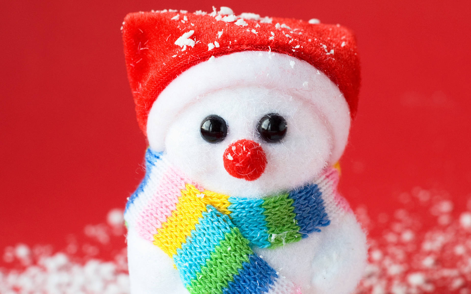 可愛的雪 桌布 壁紙 — 一個毛絨雪人的特寫，戴著紅色帽子和彩色條紋圍巾，背景是鮮豔的紅色，散落著雪花。