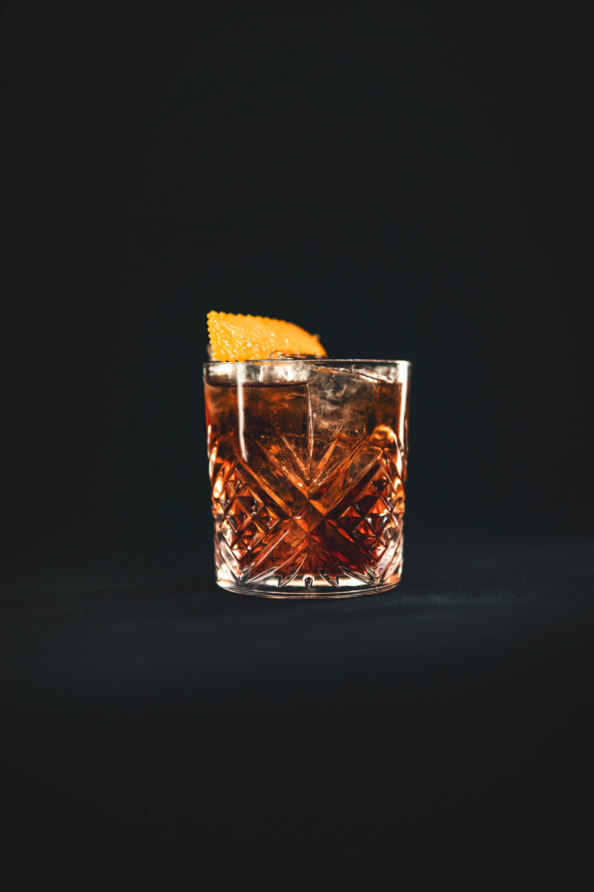Drink 桌布 壁紙 — 一個水晶切割玻璃杯，裝滿琥珀色雞尾酒和冰塊，並以橙皮裝飾，背景為黑色。